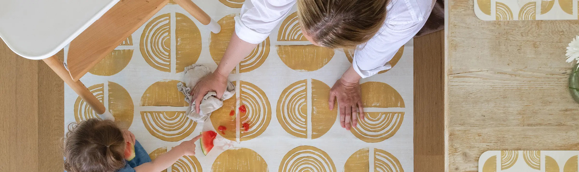 Ada Sunflower mustard yellow and white high chair mat shown from above with high chair, table, and woman wiping up a spill.
