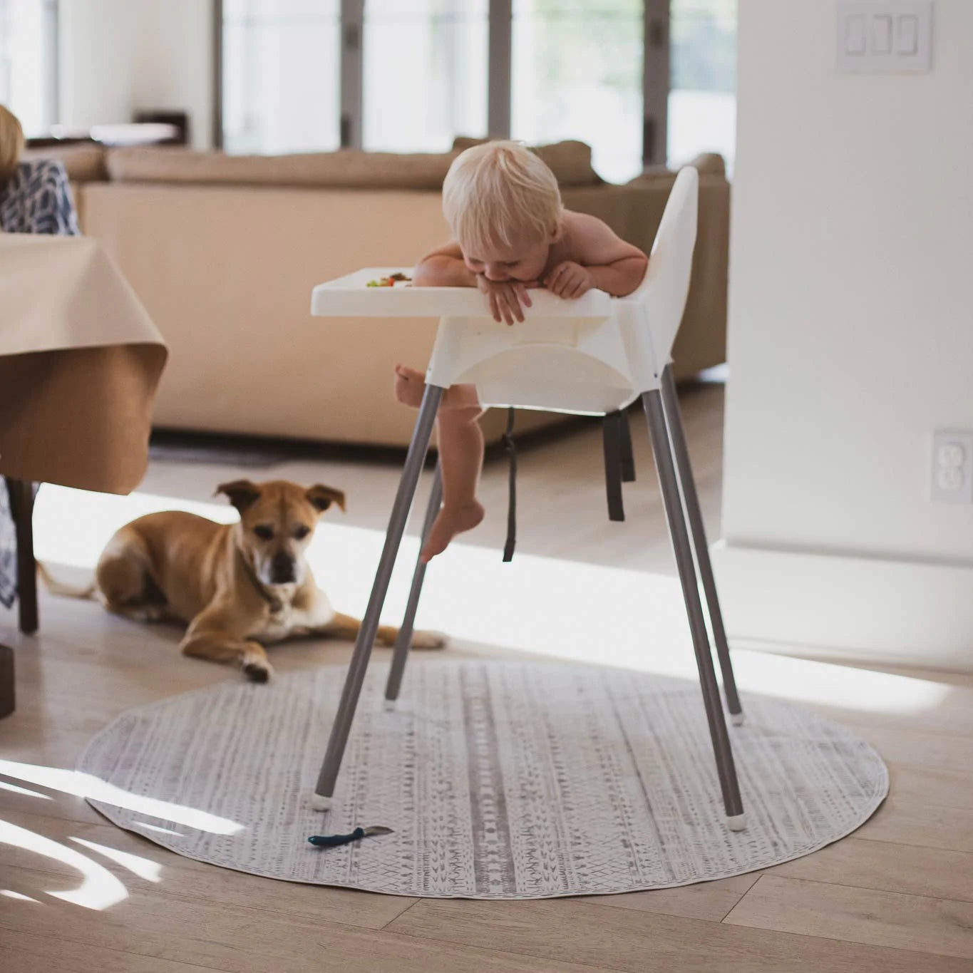 Skipping Stone gray and white striped highchair mat shown under highchair with baby laughing and dog in the background