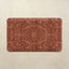 A rectangular bath mat with rounded corners featuring an intricate, symmetrical damask-style floral pattern in warm rust red and terracotta tones, displayed flat against a light beige textured background.