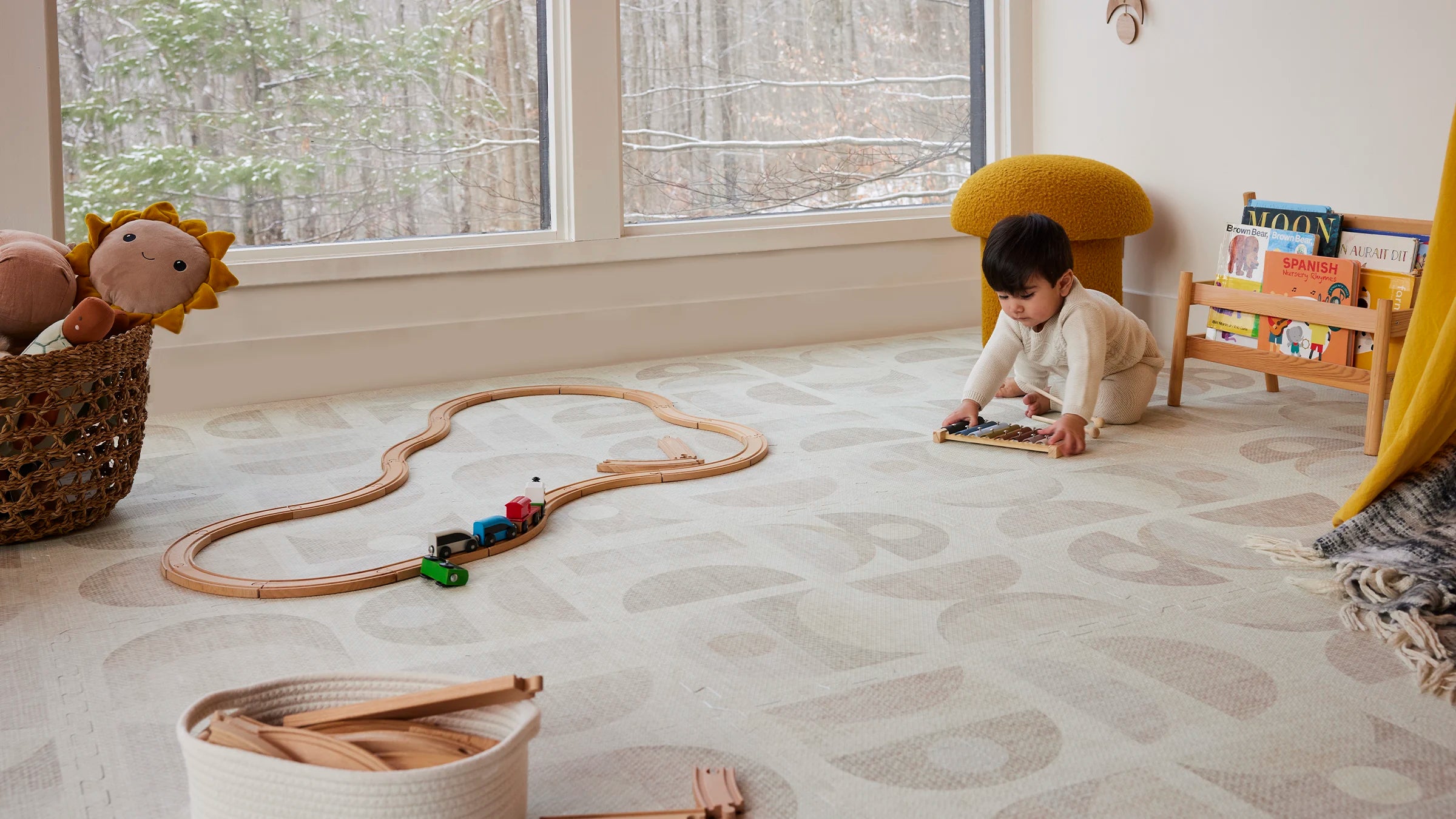 Luna sandstone play mat shown in play room with wooden train tracks, a basket of stuffed toys, a small bookshelf, and baby boy playing with xylophone  