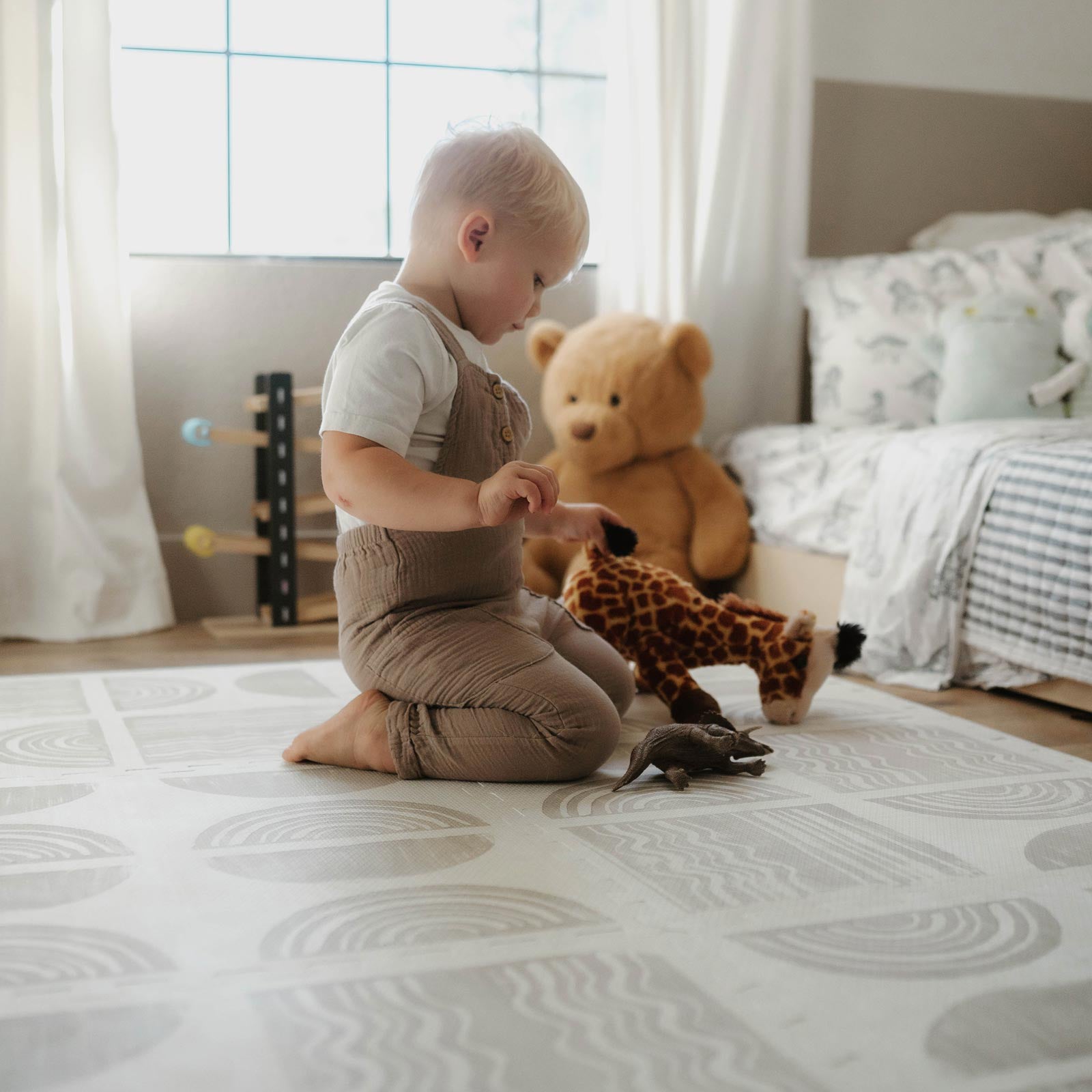 Ada Pebble beige and white block print little nomad play mat shown in bedroom with toddler boy sitting on the mat playing with  stuffed giraffe and dinosaur toy