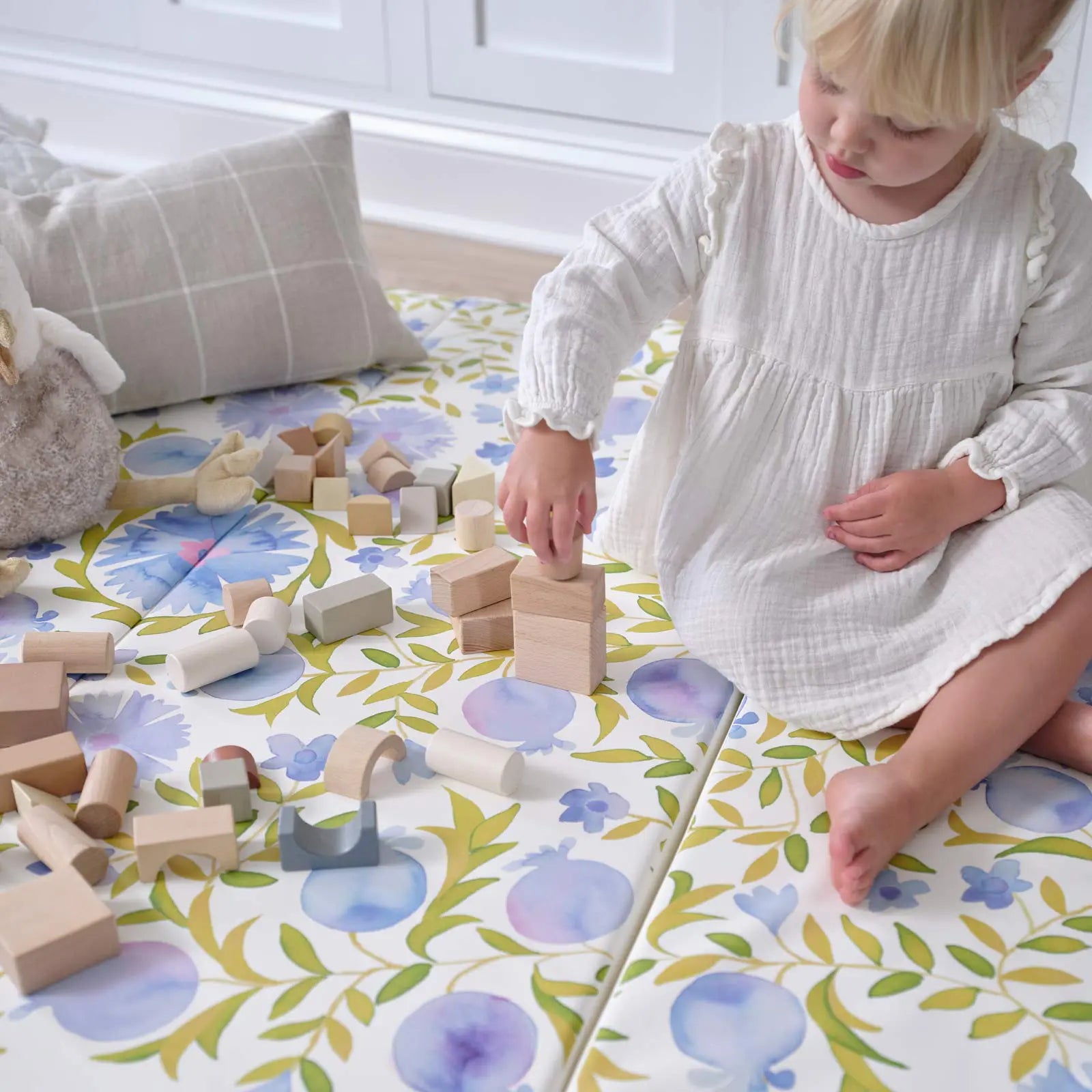 Bluebell blue green and white floral tumbling mat with toddler girl sitting on the mat next to pillows and a plush toy playing with wooden blocks
