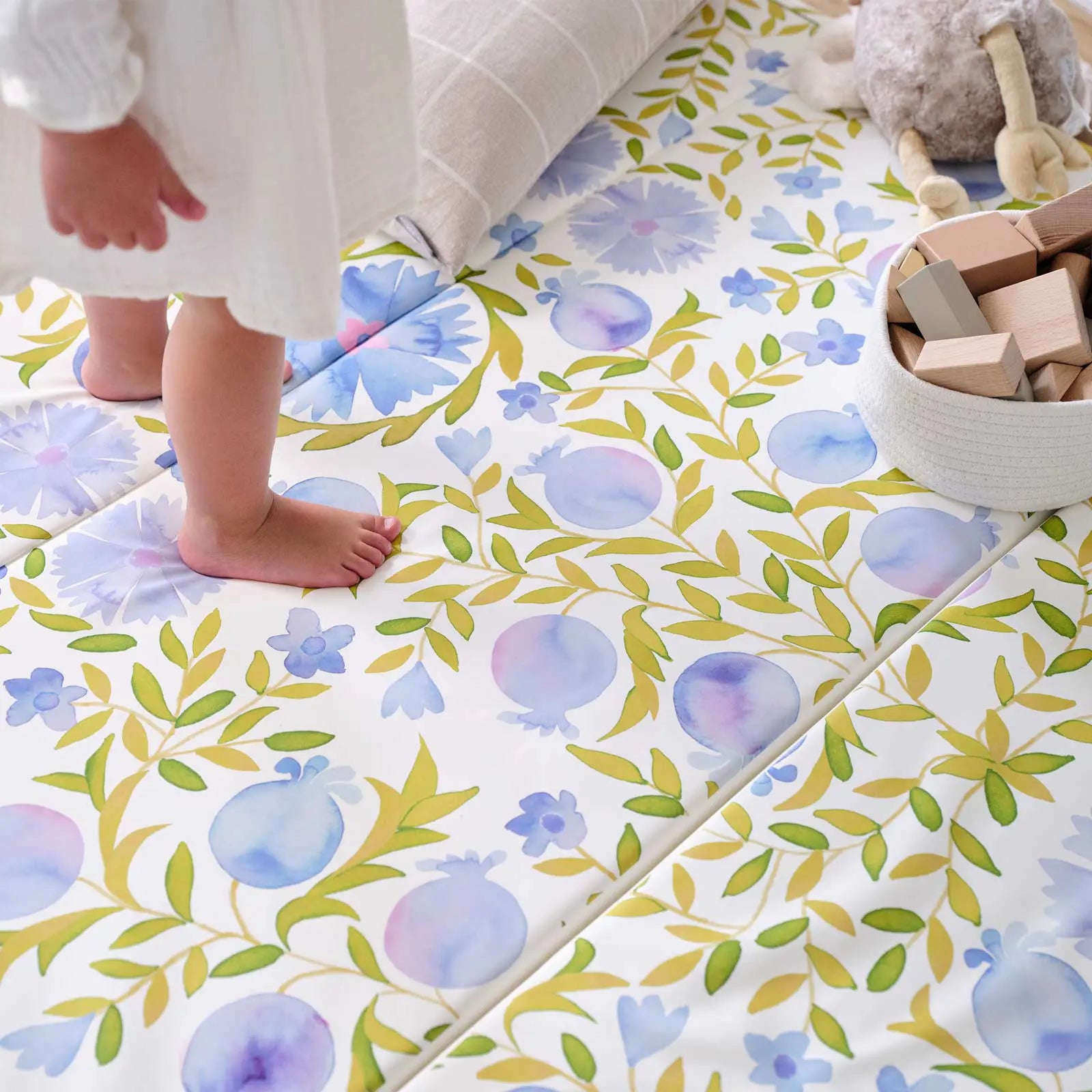 Up close shot of the Bluebell blue green and white floral tumbling mat with toddler girl feet standing on the mat and pillow, plush toy, and wooden blocks on the mat in the corner of the shot