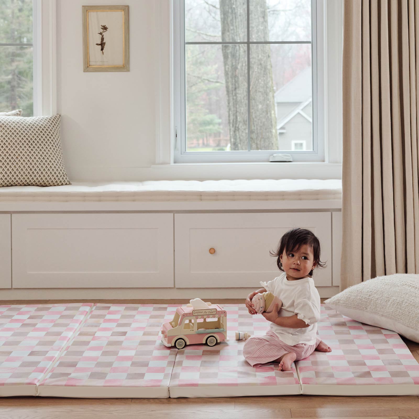 Pink and brown gingham print tumbling mat in the color Neapolitan shown in living room with toddler girl playing with ice cream truck toy on the mat