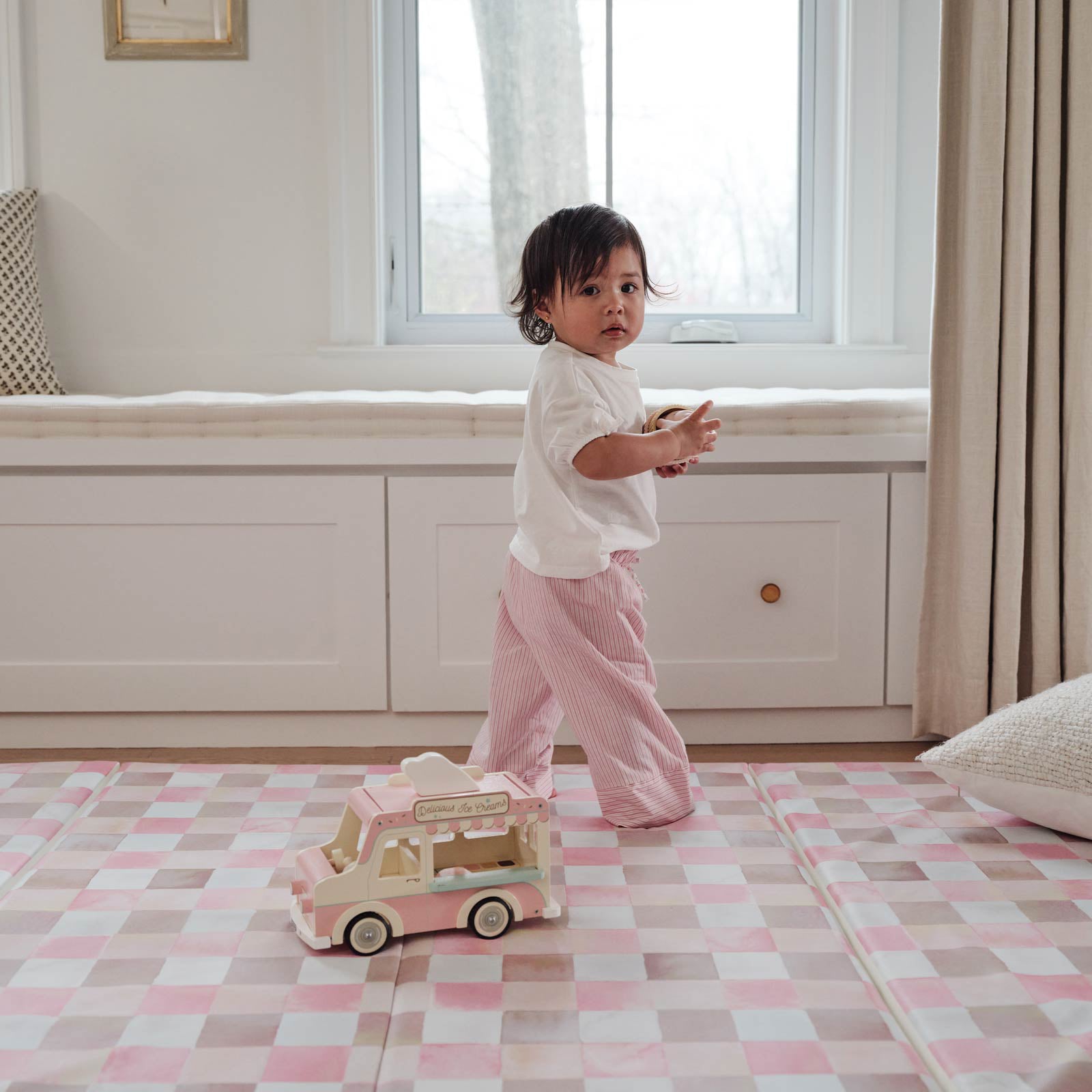 Toddler girl standing on the pink and brown gingham print tumbling mat holding an ice cream toy