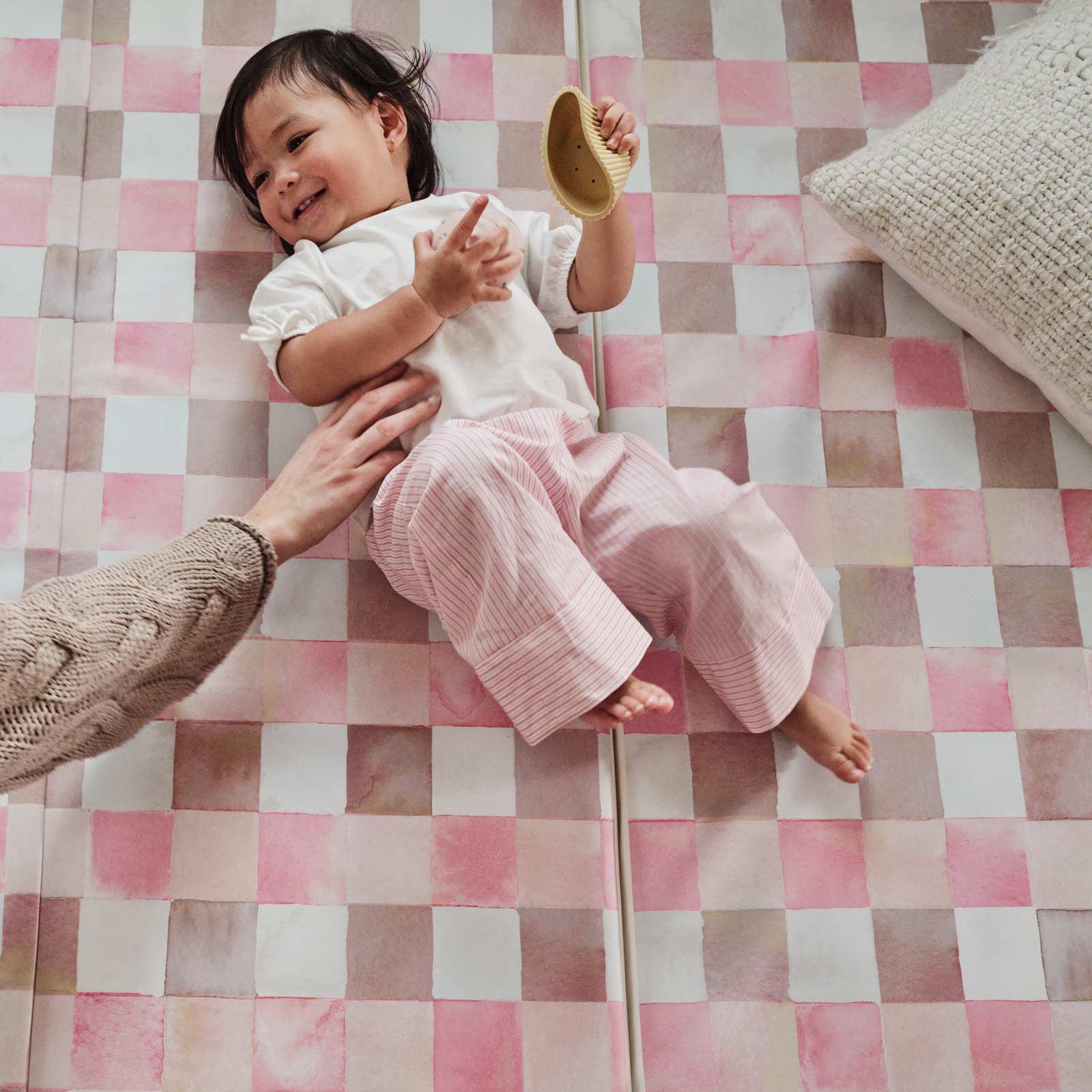 Neapolitan pink and brown gingham print tumbling mat shown with toddler girl laying on the mat playing with a toy being tickled