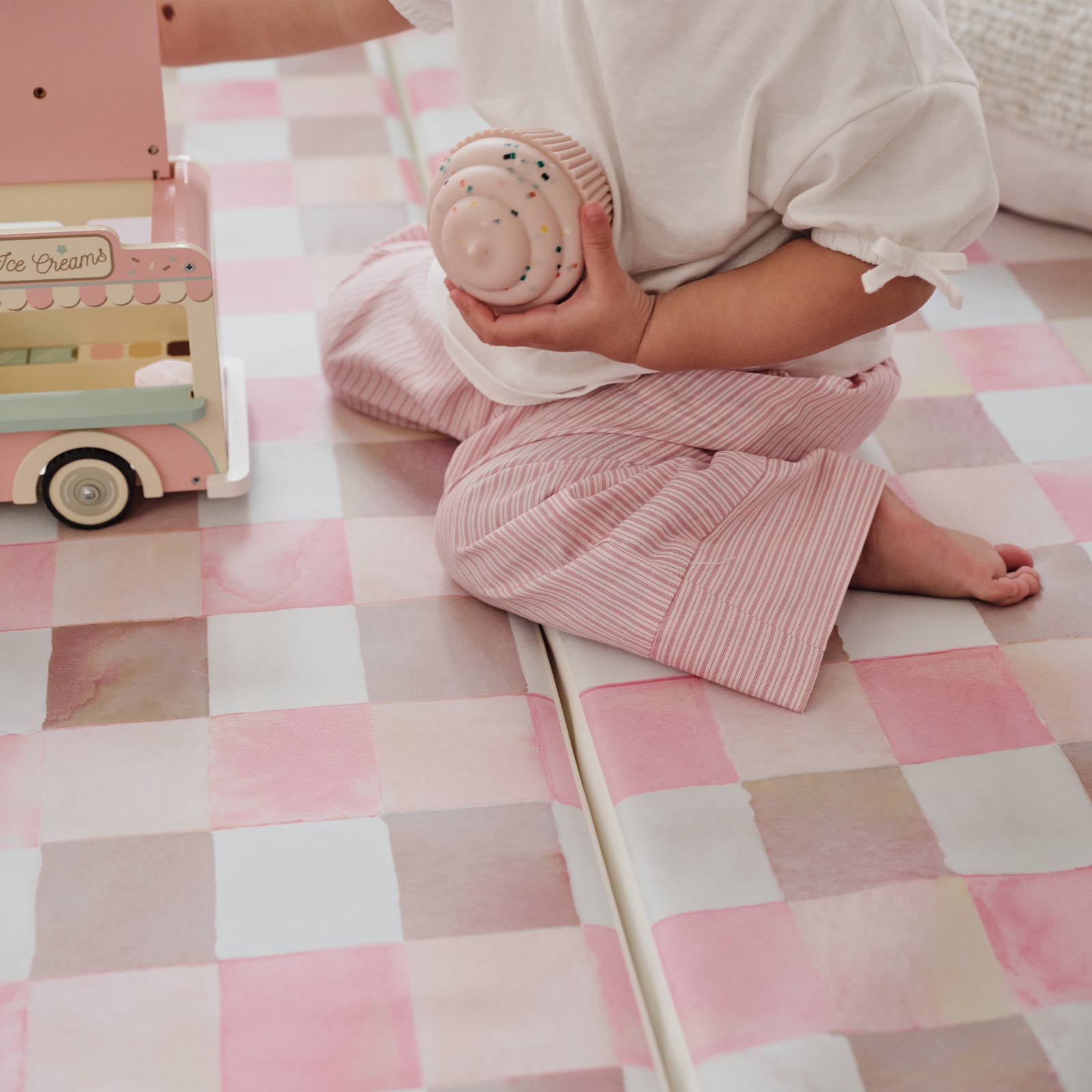 Close up shot of a toddler girl playing with an ice cream truck toy sitting on the pink and brown gingham print neapolitan tumbling mat. 