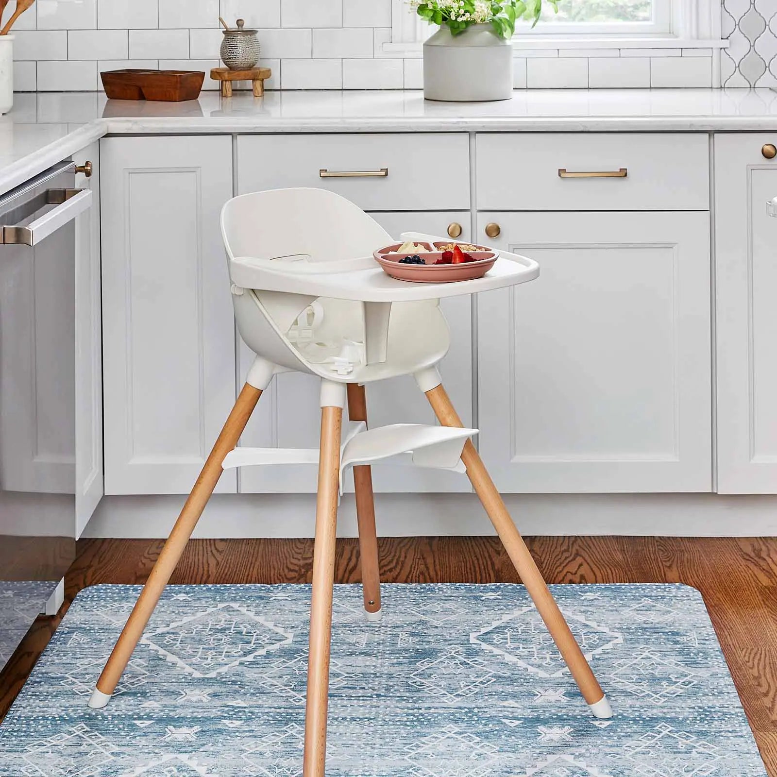 Ula indigo blue and white boho print highchair mat shown in a kitchen under a highchair with a plate of fruit on it