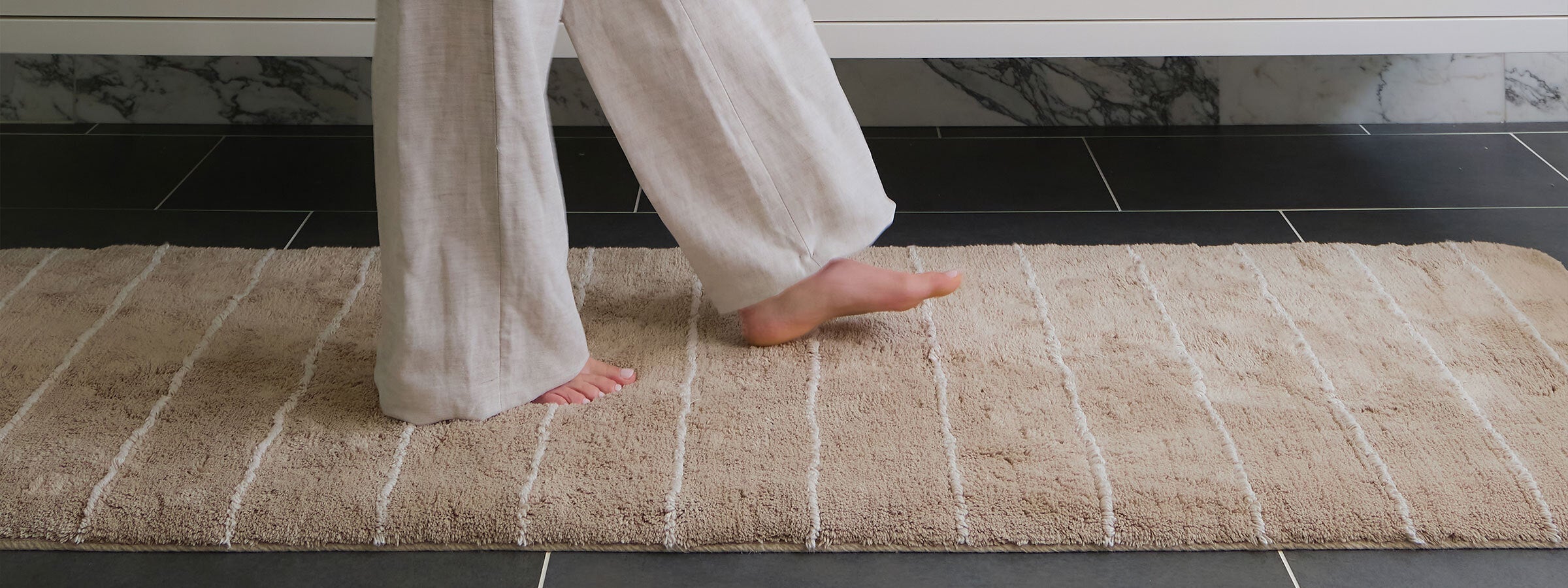 Ula driftwood neutral boho standing mat shown with womans feet standing on the mat in front of a kitchen counter on a wood floor