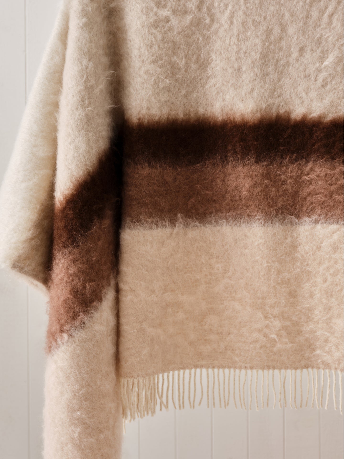 Beige and brown striped mohair blanket with fringes in front of a white wall.