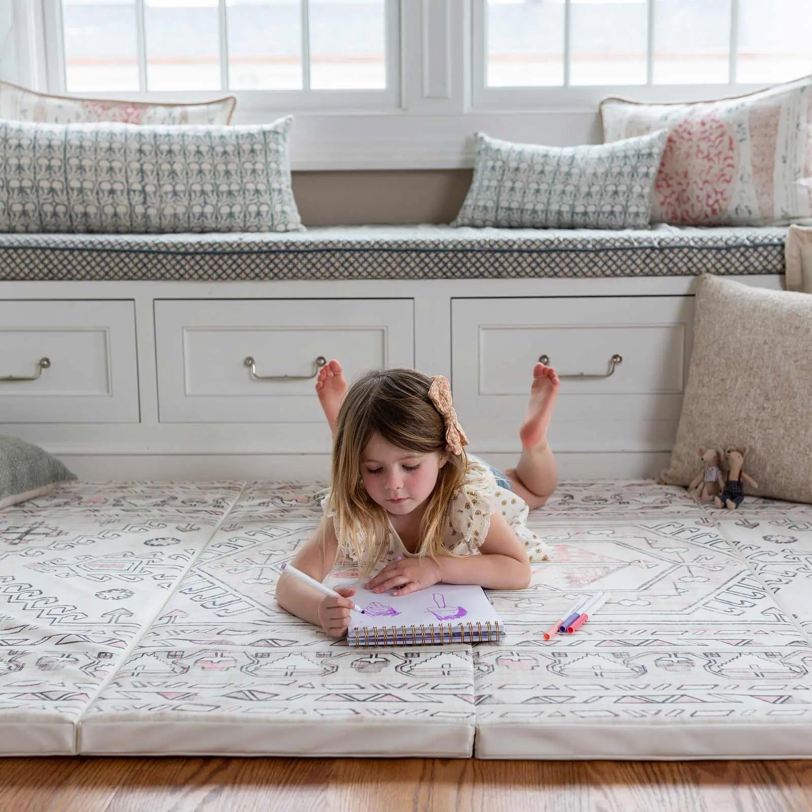 Aryla Shifting Sands cream and pink boho tumbling mat shown in front of a window seat with little girl coloring while laying on the mat