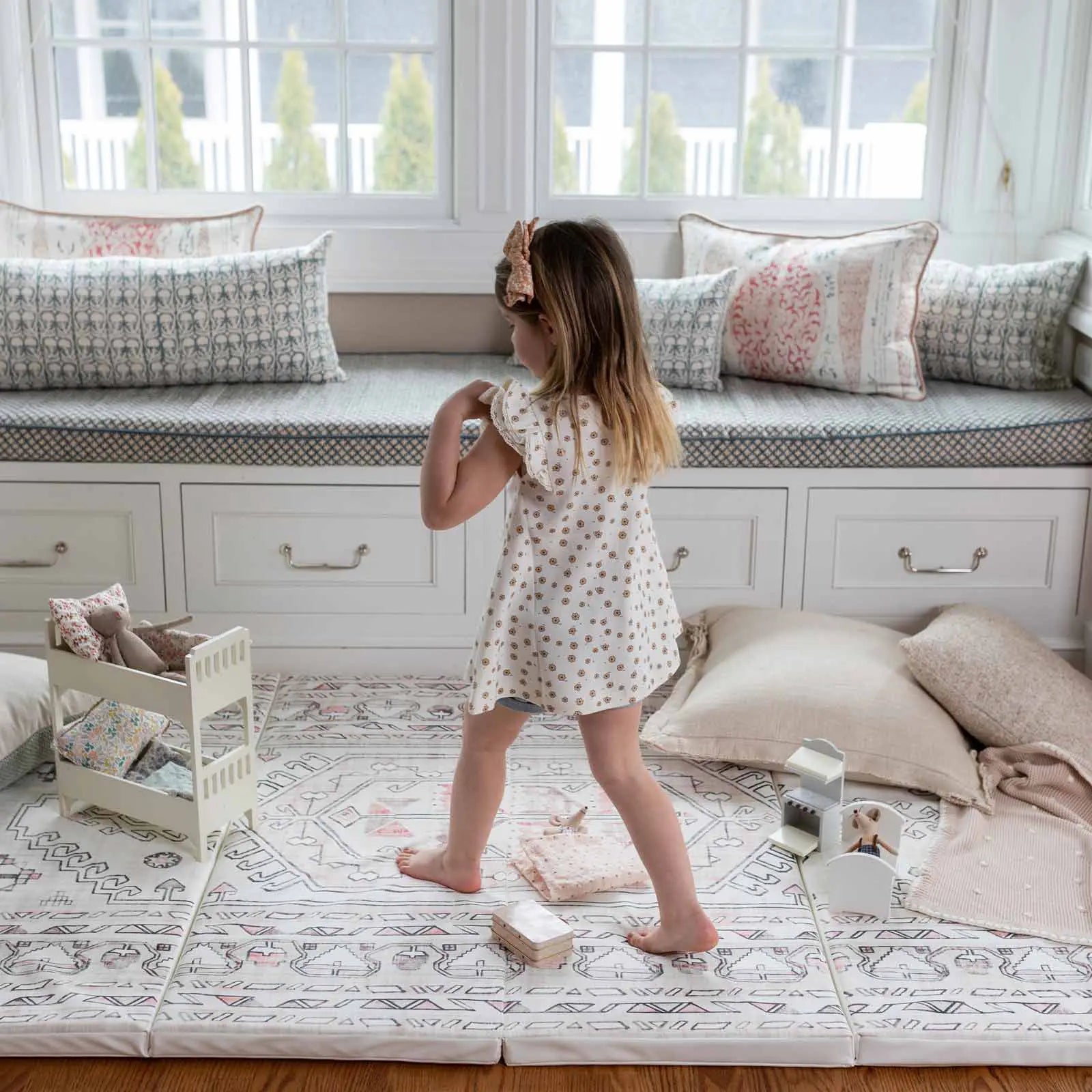 Aryla Shifting Sands cream and pink boho tumbling mat shown in front of a window seat with pillow and toys and little girl walking on the mat