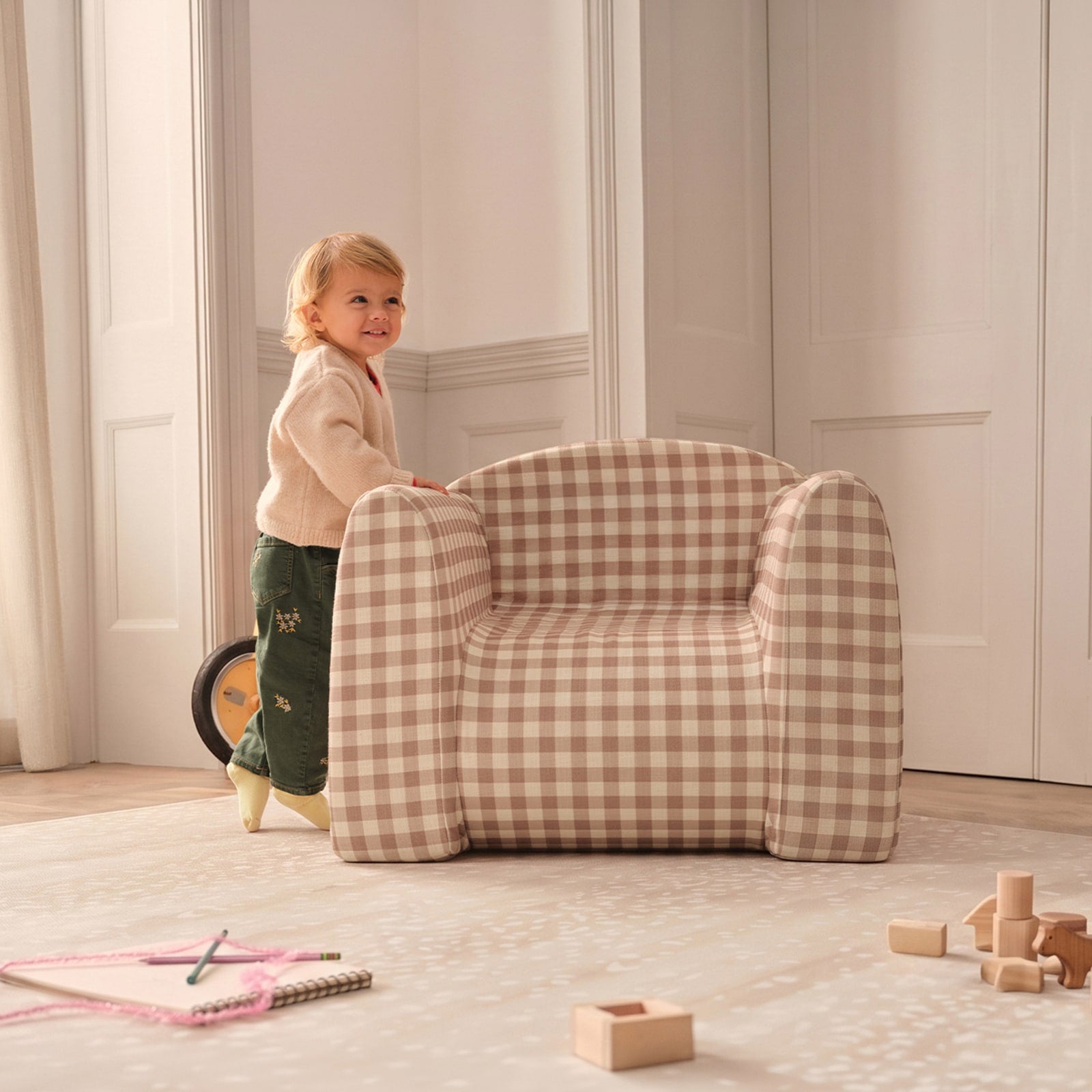 A toddler stands beside a small upholstered armchair in a softly lit room. The chair has a beige and white gingham check pattern.