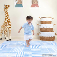 hadley french blue inverted stripe blue and white tumbling mat shown in a plat room with stacked baskets and giant giraffe plush toy with toddler boy running across the mat. @anaperu written in the lower left hand corner
