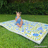 suzette bluebell blue and green floral tumbling mat shown laying on grass outside with toddler girl sitting on the mat eating a snack. @charlicevans written in the lower right hand corner.