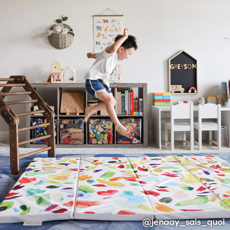 confetti multi rainbow brushstroke pattern tumbling mat shown in play room with little boy jumping off wooden playspace onto mat. @jenaay_sais_quoi written in lower right hand corner.