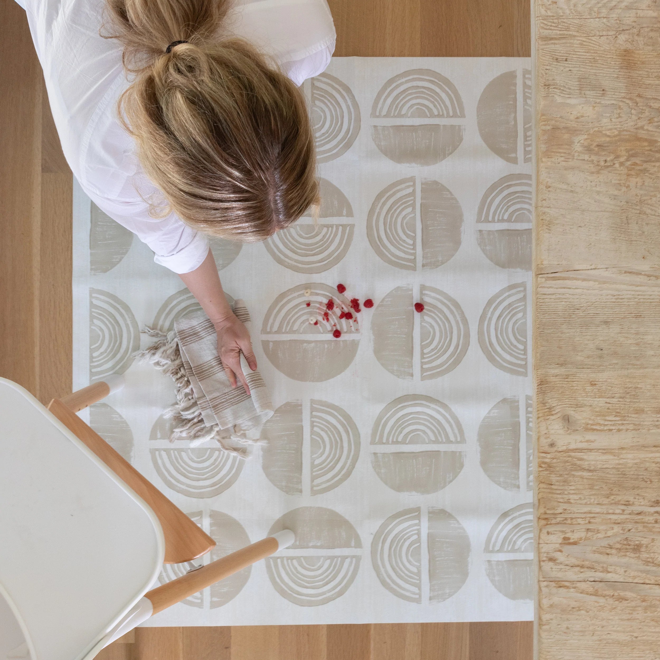 Ada modern minimalist baby high chair mat in Pebble taupe and off white. Shown from above with woman cleaning up a spill under a high chair.