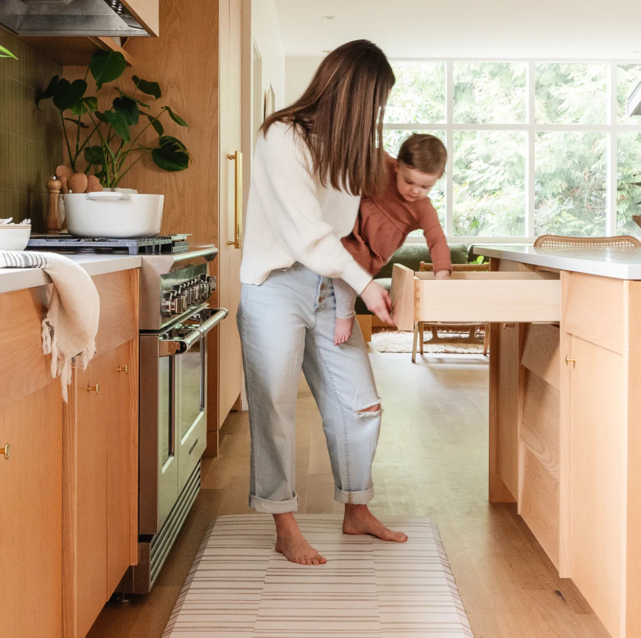 Nara natural beige and white inverted stripe kitchen mat with woman holding a baby standing on the mat in a kitchen.