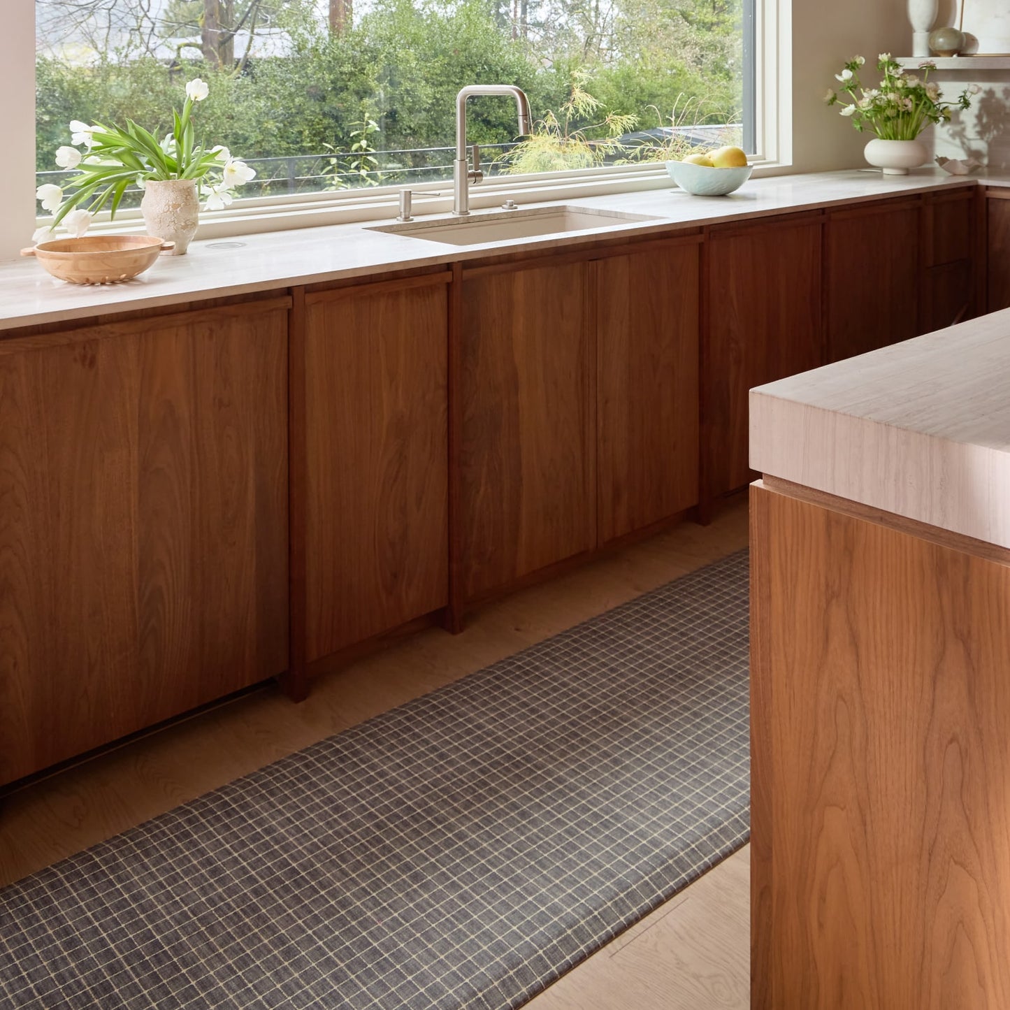 Modern kitchen with wooden cabinets, a sink, grid pattern kitchen mat, and a window view of greenery.