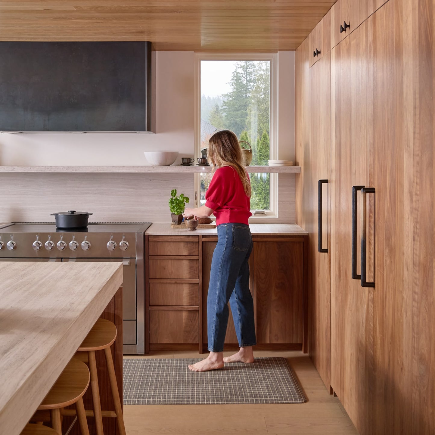 Woman in a modern kitchen with wooden cabinets and stainless steel appliances standing on a brown kitchen mat.