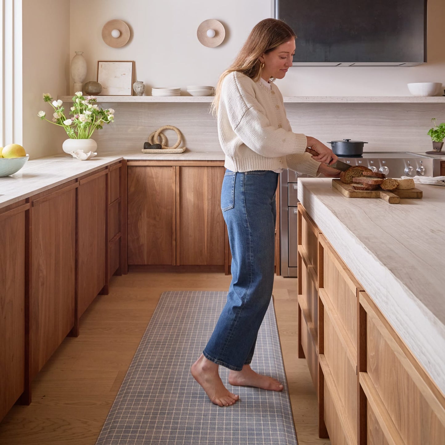 Woman standing in a modern kitchen with wooden cabinets and a grid pattern kitchen rug.