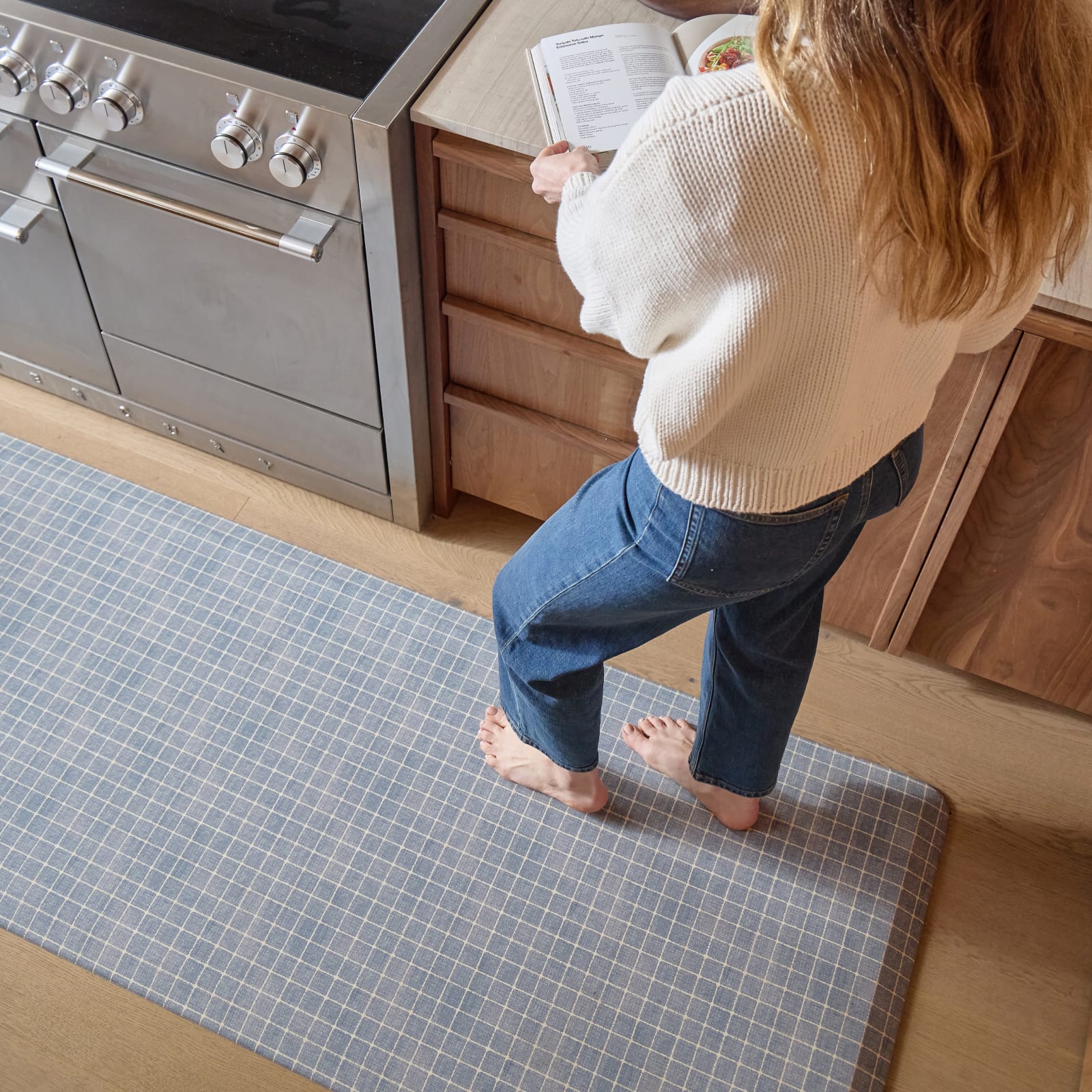 Person standing on a grid pattern mat in a kitchen, wearing a beige sweater and blue jeans.