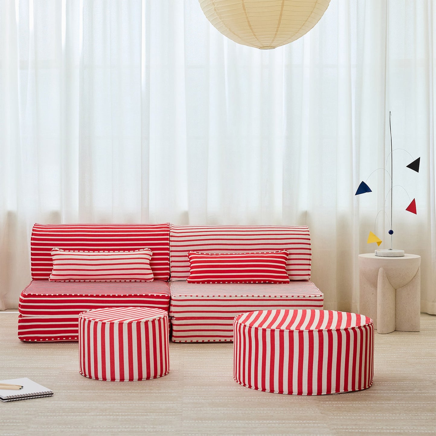 A modern living room setup featuring a red and white striped modular sofa, two matching striped ottomans, sheer white curtains, and a light wood floor.