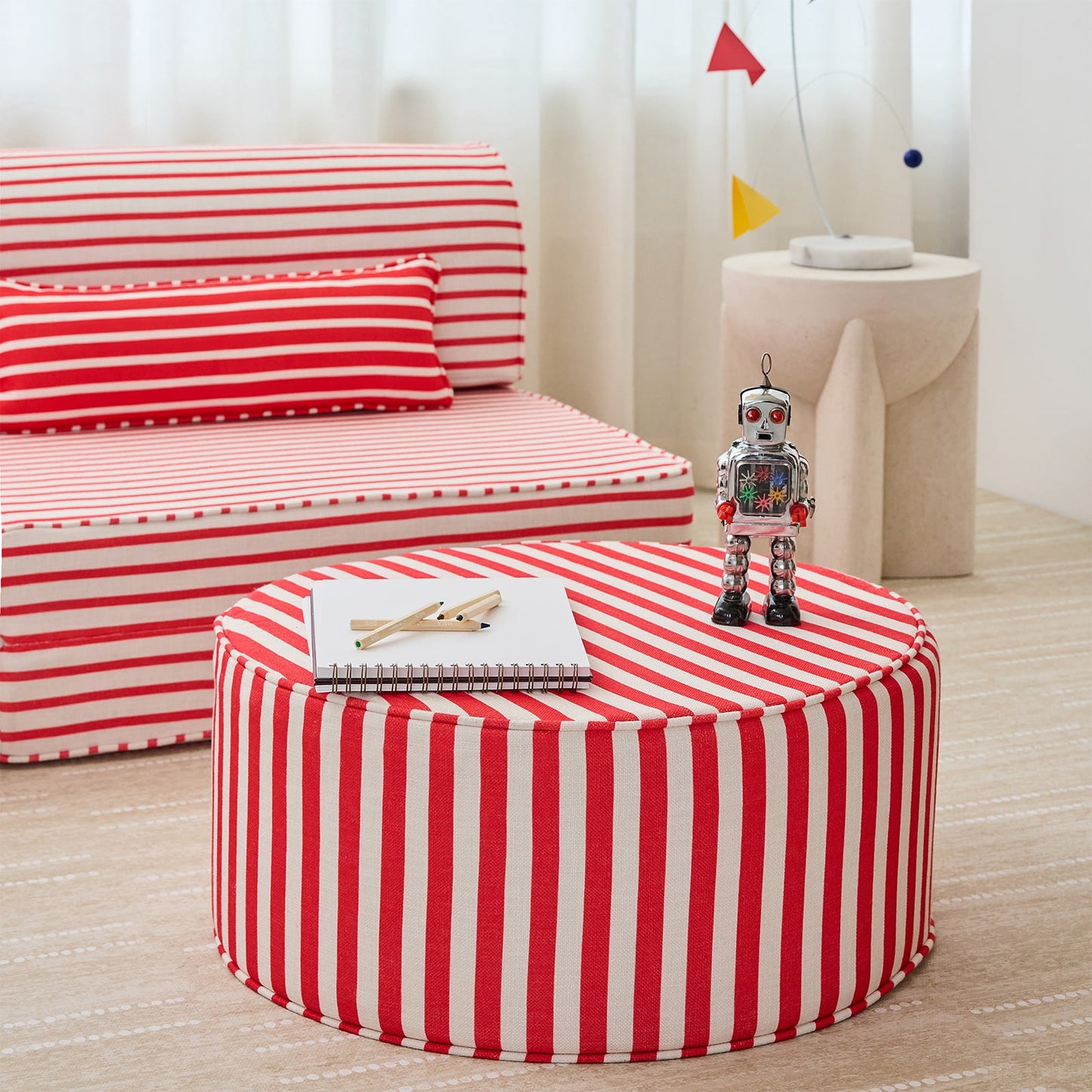 A round ottoman or pouf in front of a matching striped sofa, both upholstered in a red and white vertically striped fabric. The scene is styled in a child's room with a small toy robot on top of the ottoman, a notebook, and a side table in the background.