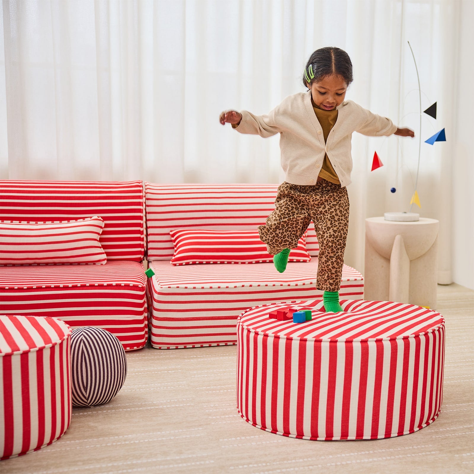 A child stands on a round red-and-white striped ottoman in a bright, modern playroom. The room features matching red-and-white striped furniture, including a sofa, pillows, and another ottoman in the foreground, with a light wood floor and white curtains in the background.