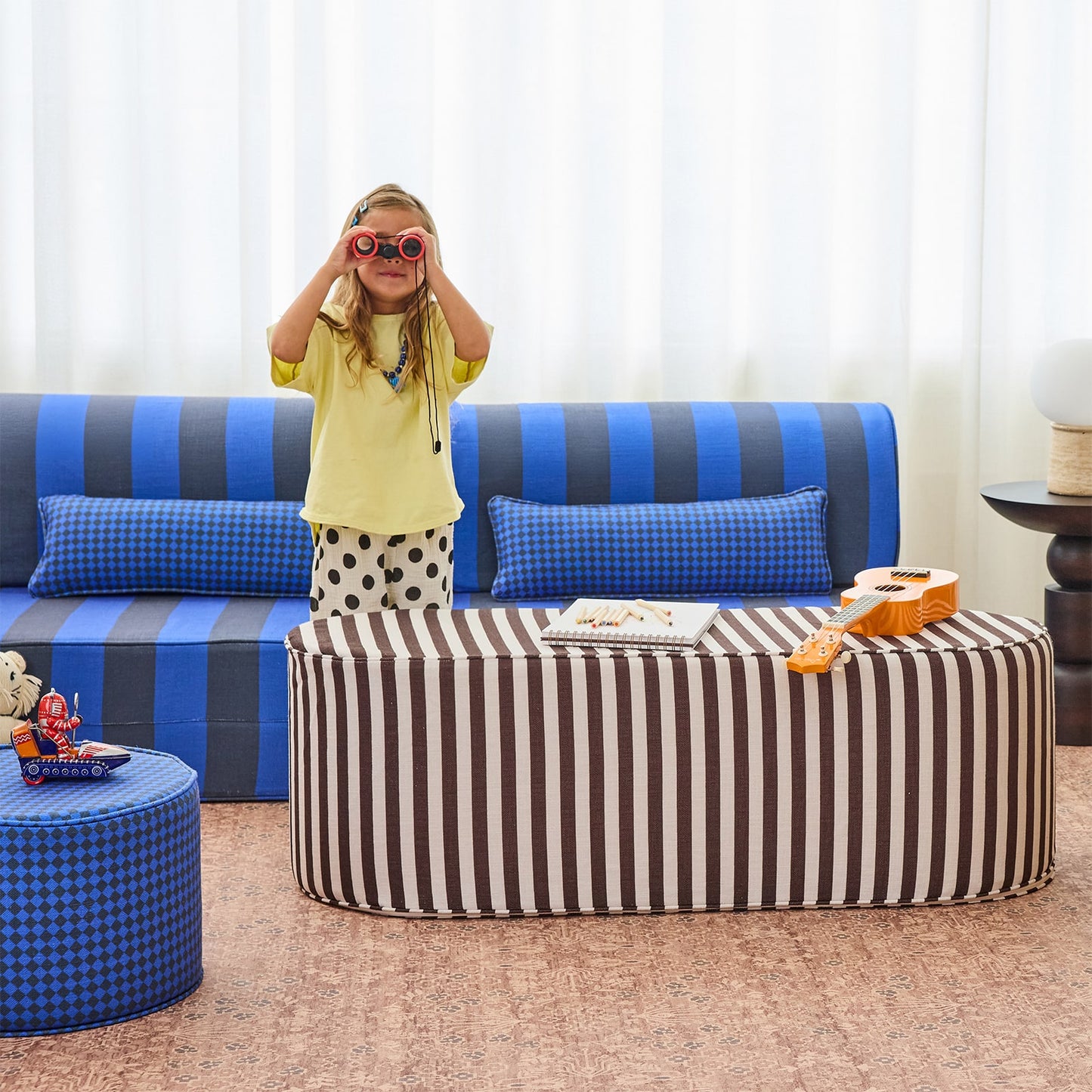 A child stands in front of a blue and gray striped sofa wearing a yellow shirt, polka-dot shorts, and red sunglasses, with a matching striped ottoman and blue pillows in a bright living room setting.