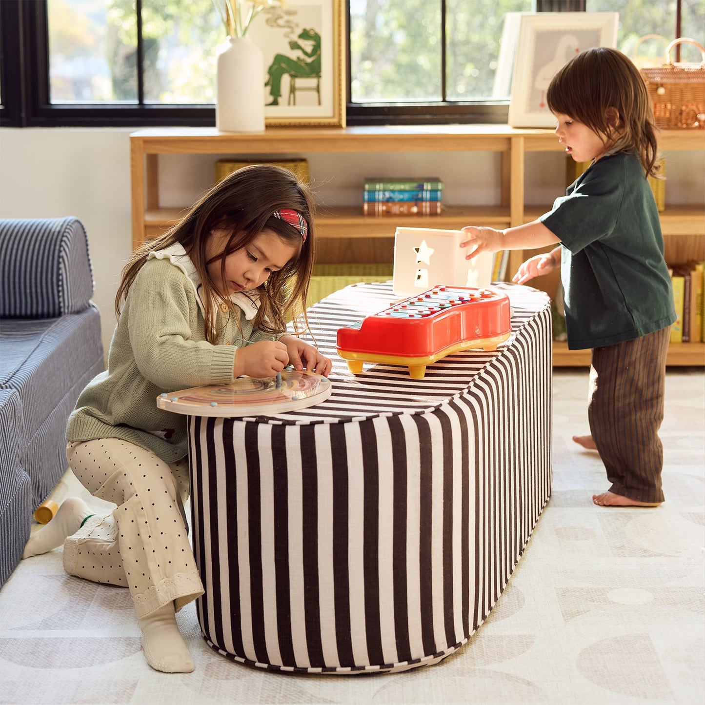 A child's coffee table with a black and white striped fabric cover, set in a bright living room. A young girl sits on the floor playing with a toy, while a boy stands nearby holding a toy phone.