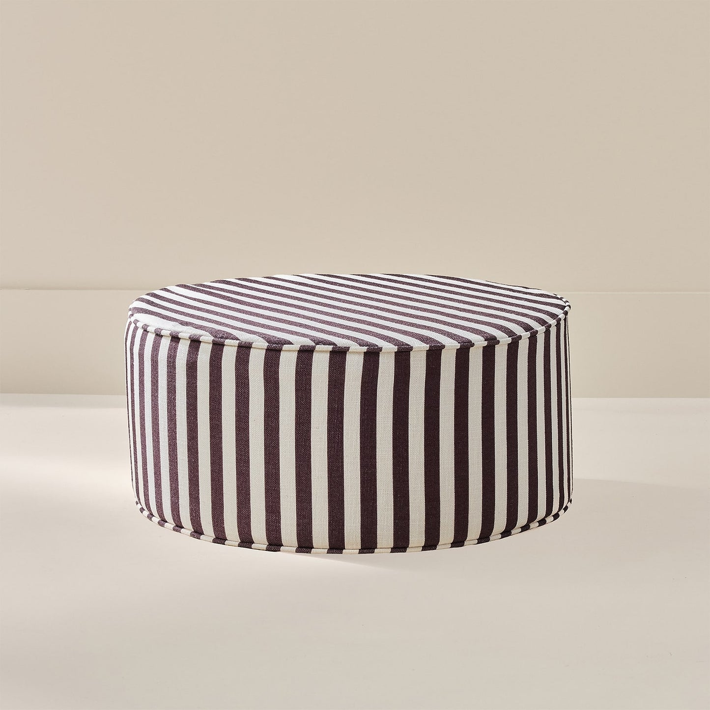 A round upholstered ottoman with vertical dark brown and white stripes, shown on a clean beige studio background.