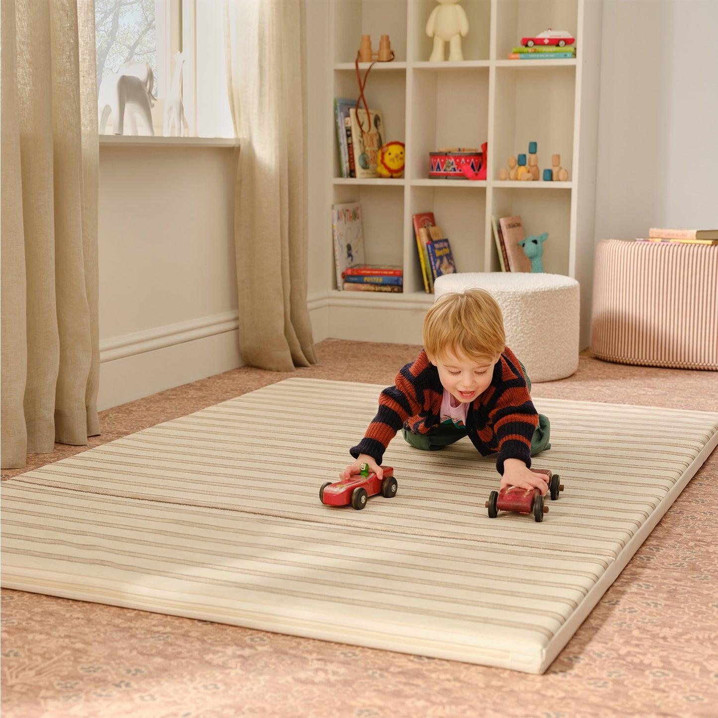 Child playing with toy cars on a striped mat in a room with shelves and toys.