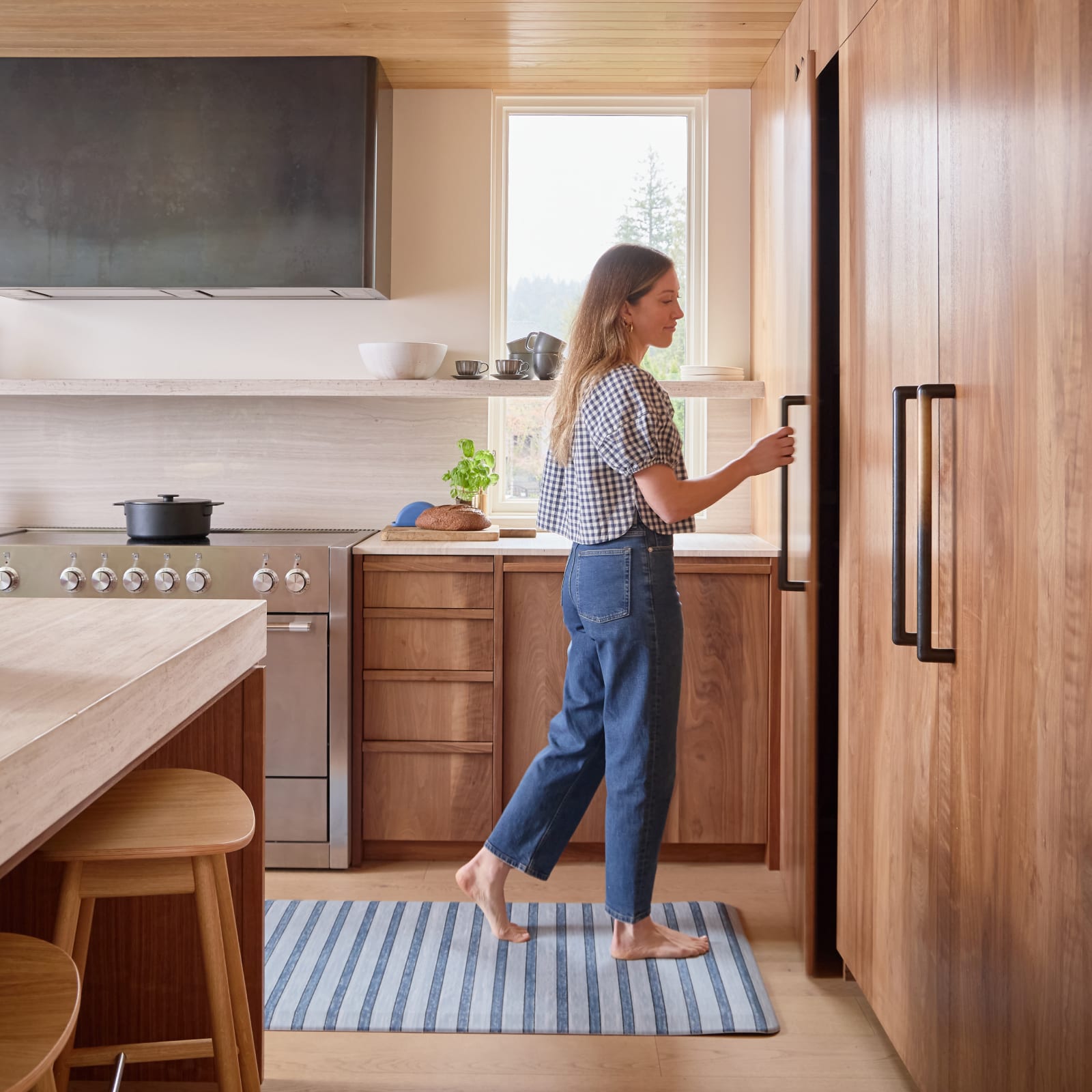 Woman standing in a modern kitchen with wooden cabinets and a striped kitchen mat.