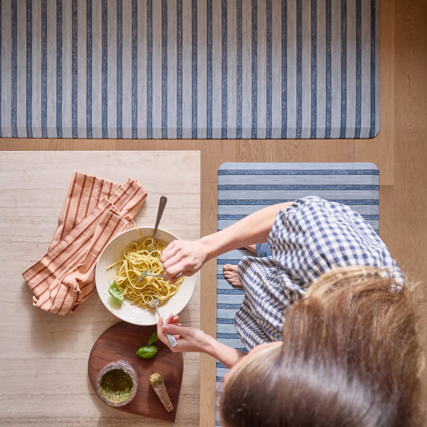Woman standing at a kitchen counter with a plate of pasta standing on a striped blue rug.