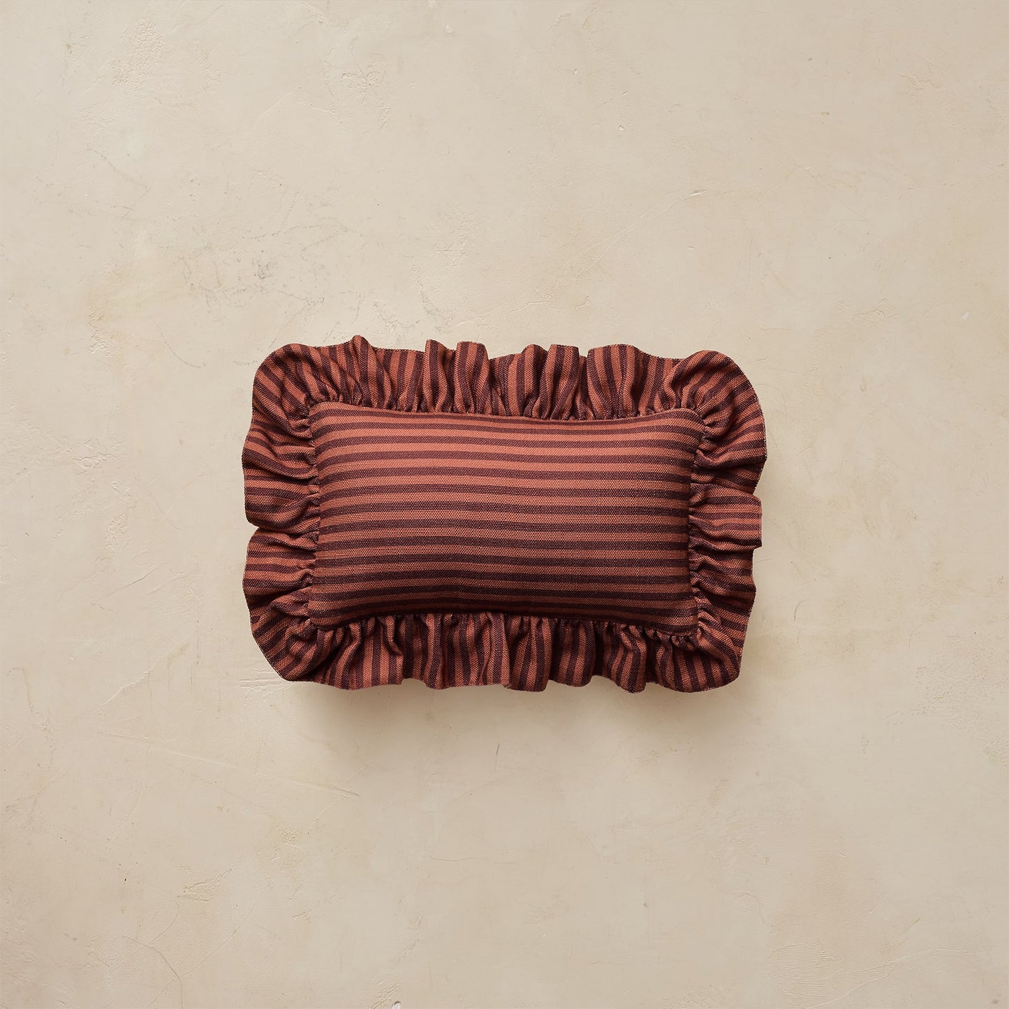 A rectangular decorative pillow with ruffled edges, featuring horizontal stripes in rust red and maroon, set against a textured beige background.