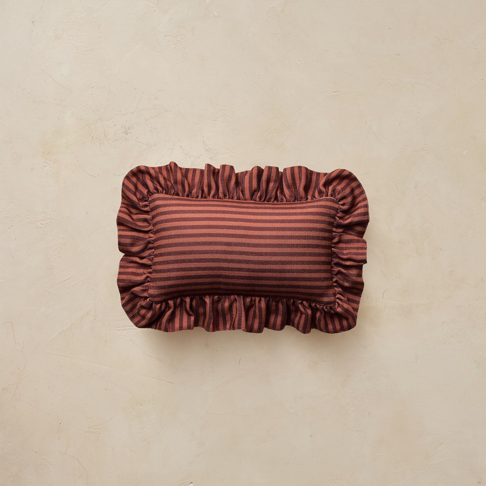 A rectangular decorative pillow with ruffled edges, featuring horizontal stripes in rust red and maroon, set against a textured beige background.