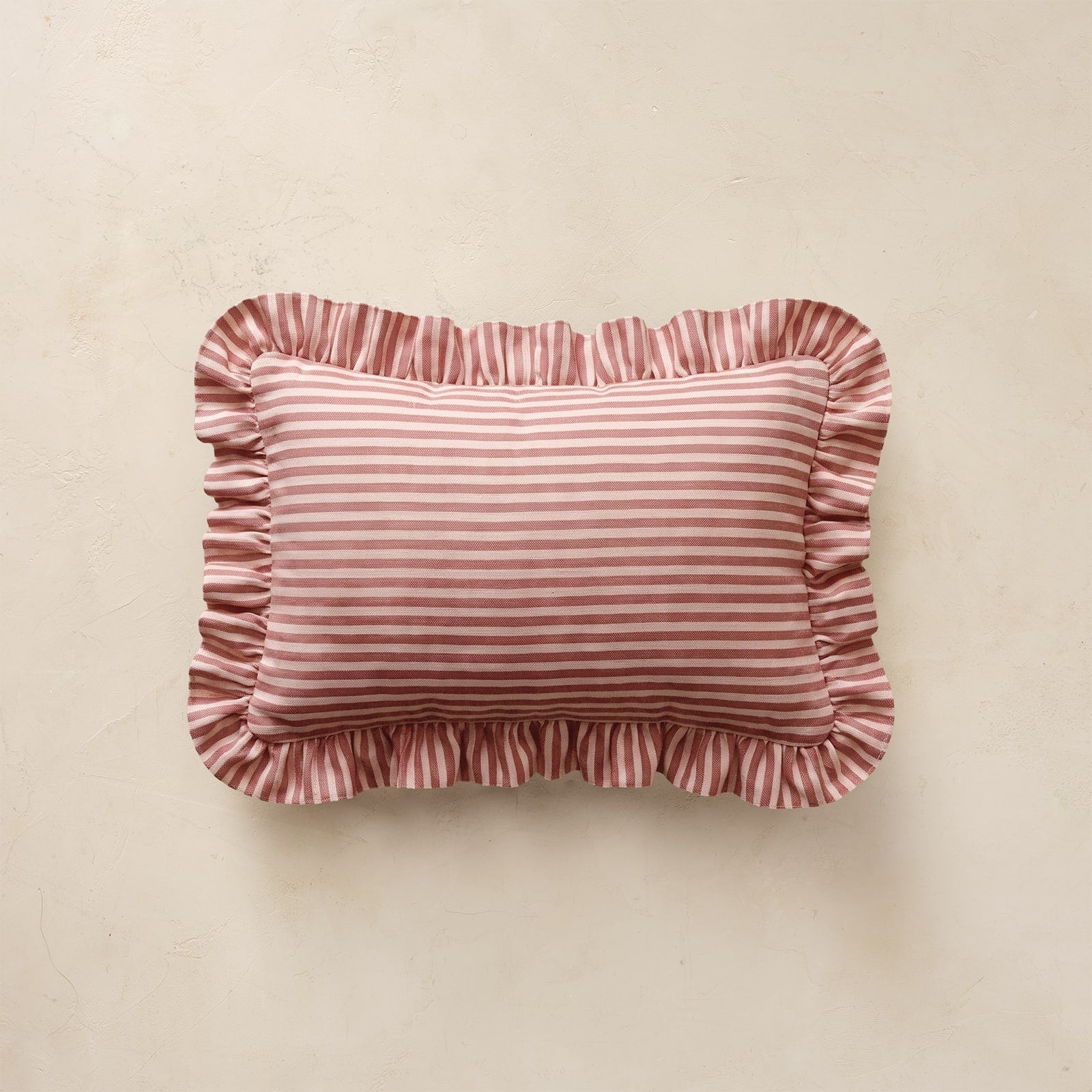 A rectangular decorative pillow with ruffled edges, featuring a dusty pink  horizontal stripe pattern, displayed against a textured beige background.