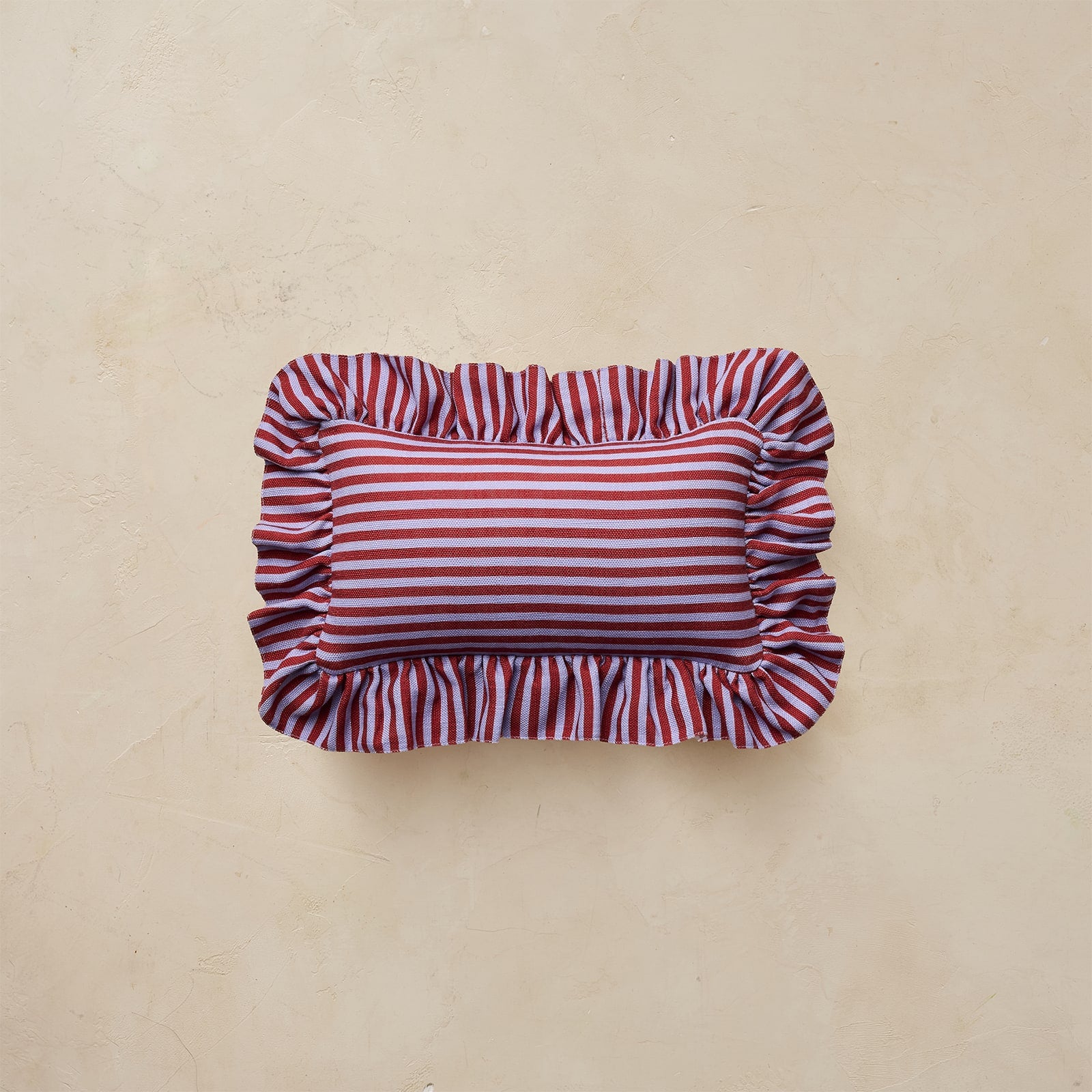 A rectangular throw pillow with red and white horizontal stripes and ruffled edges, displayed on a textured beige background.