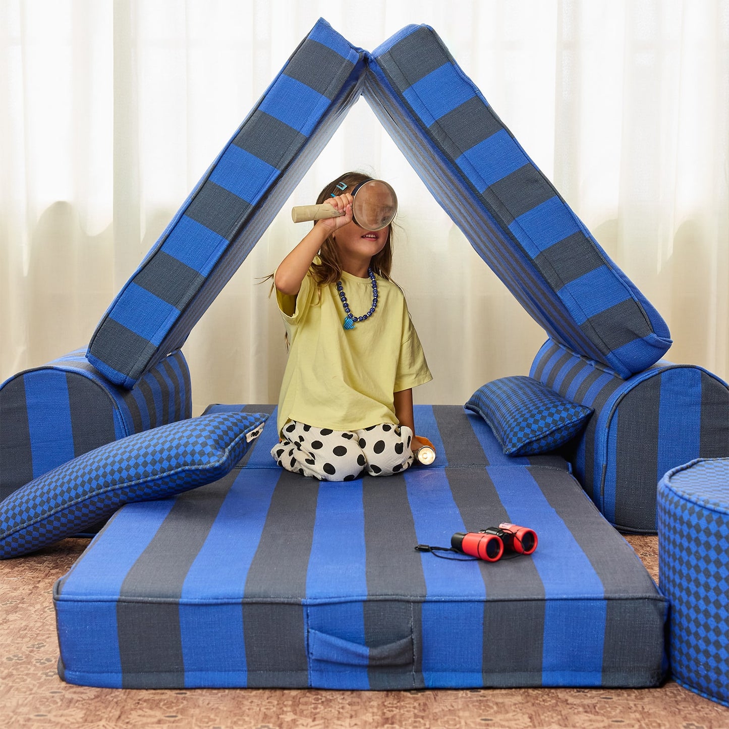 A child sits inside a blue and gray striped foam play fort with matching pillows, using toy binoculars, in a brightly lit indoor room.