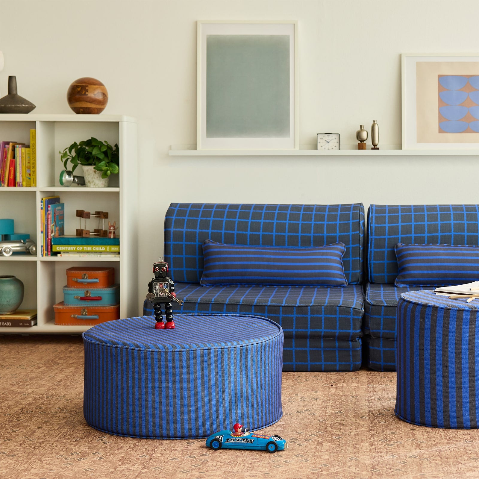 A styled children's room scene featuring a blue and black plaid sofa, a matching striped ottoman table, and a striped side table on a beige carpet against a white wall with framed art.