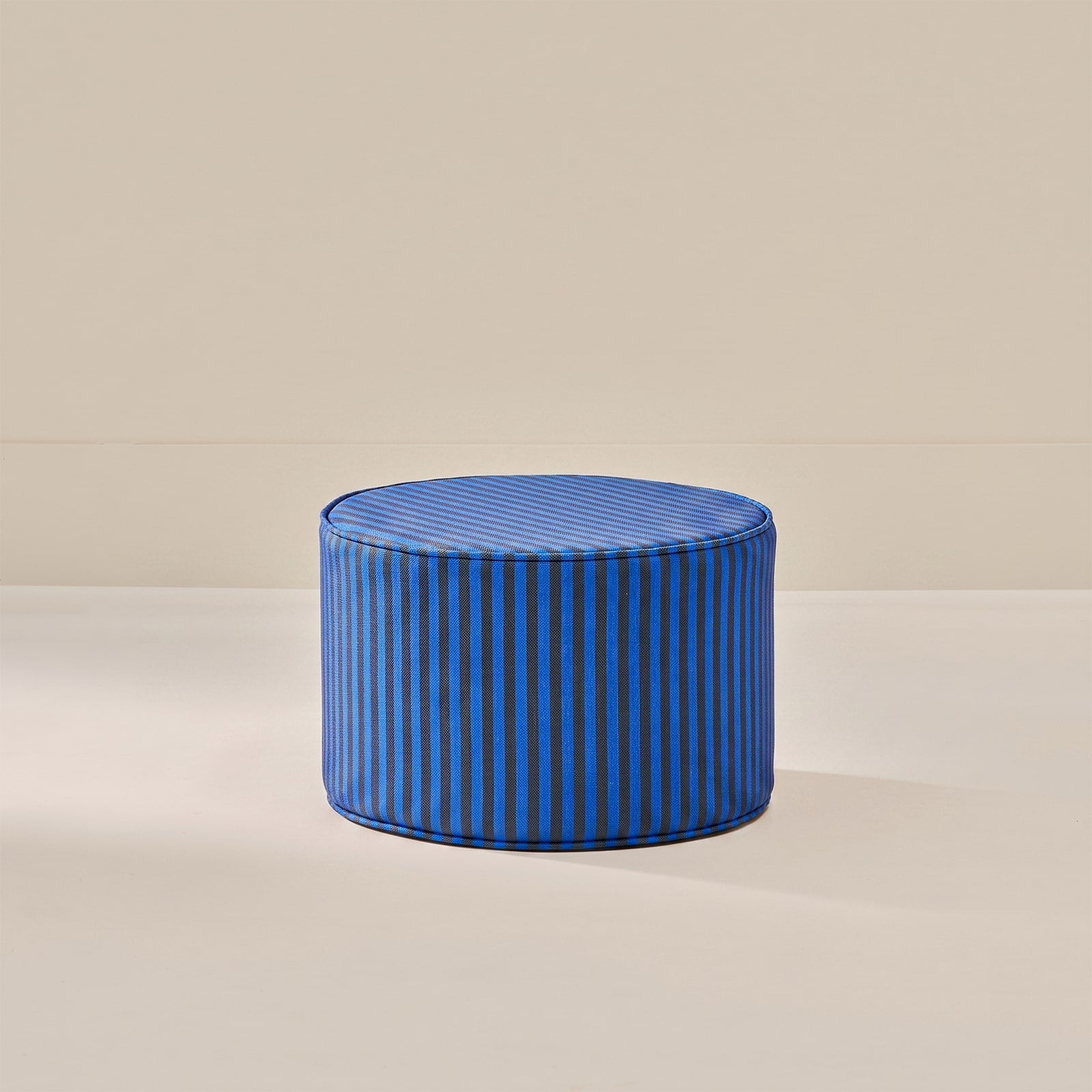 A round blue pouf ottoman with vertical stripes, set against a minimalist beige background.