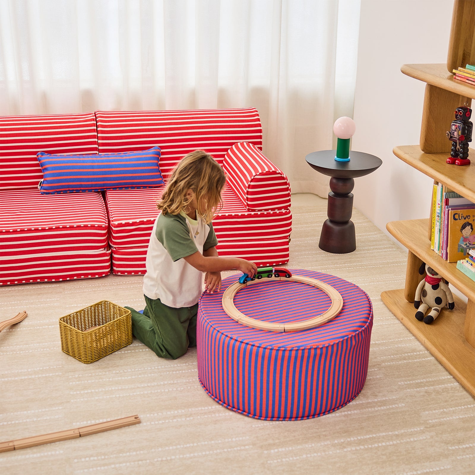 A child kneels on a light wood floor in a bright room, playing with a small toy train on a round striped ottoman. A red-and-white striped sofa, blue pillow, side table, and wooden bookshelf are visible in the background.