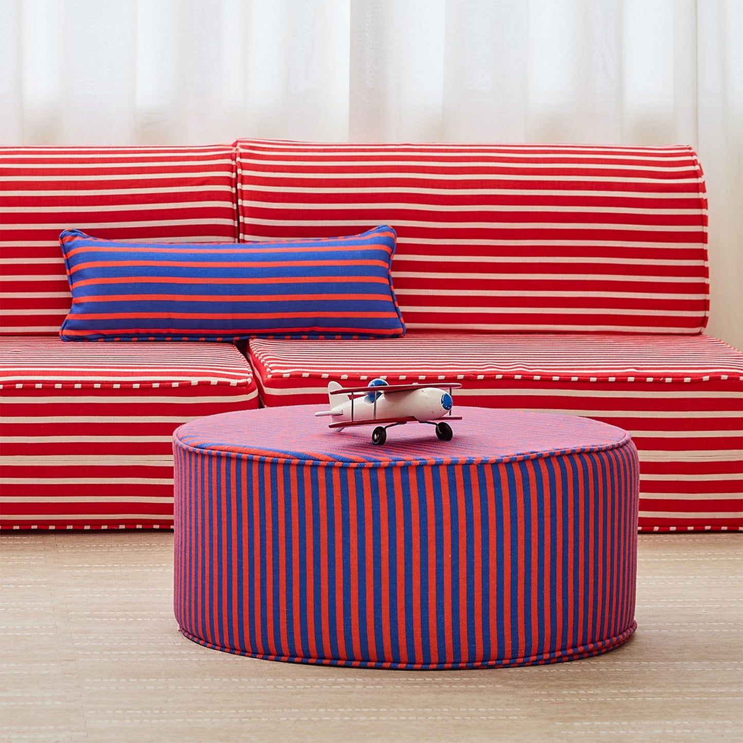 A children's play couch and ottoman set in a bright room, featuring red and white striped cushions on the couch and a round ottoman with red and blue vertical stripes in the foreground.