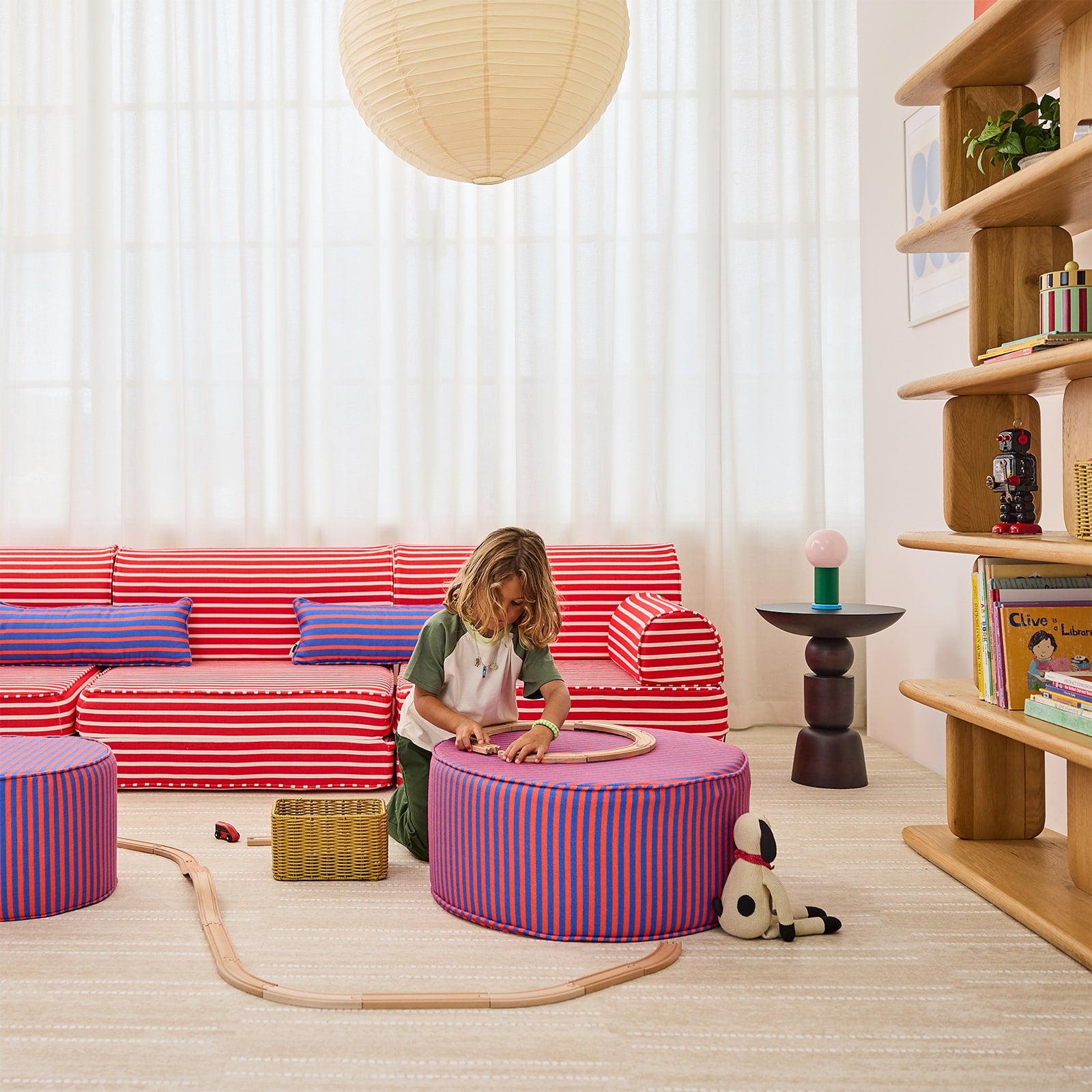 A child sits on a round striped ottoman in a bright, modern children's room. The room features a red and white striped sofa, a wooden bookshelf, a hanging paper lantern, and a light-colored rug.