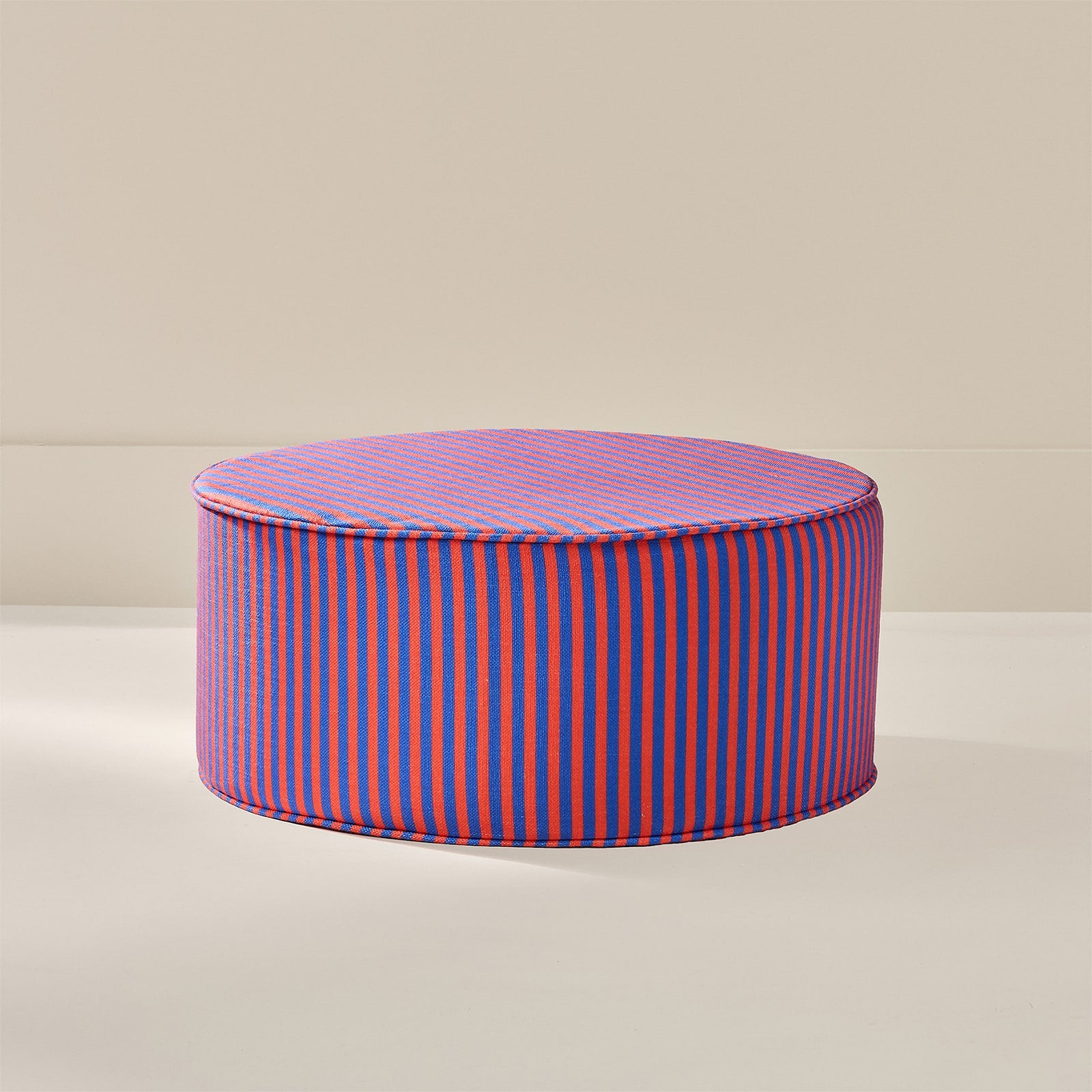 A round upholstered ottoman or pouf with a lid, featuring vertical stripes in bright blue and orange-red, set against a minimalist beige background.