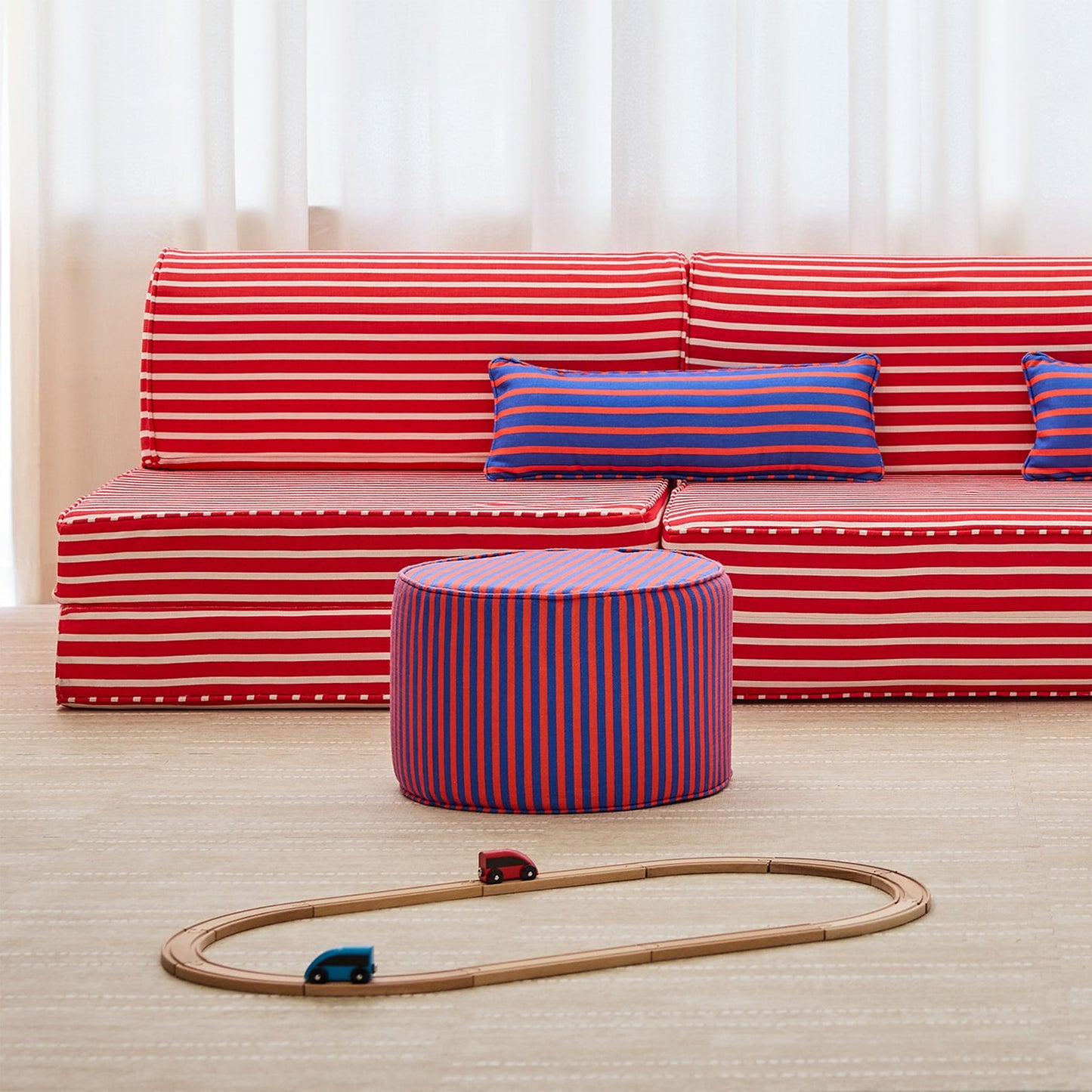 A children's play area setup featuring a red and white striped modular sofa, a matching red and white striped ottoman, and blue and red striped accent pillows on a light wood floor.