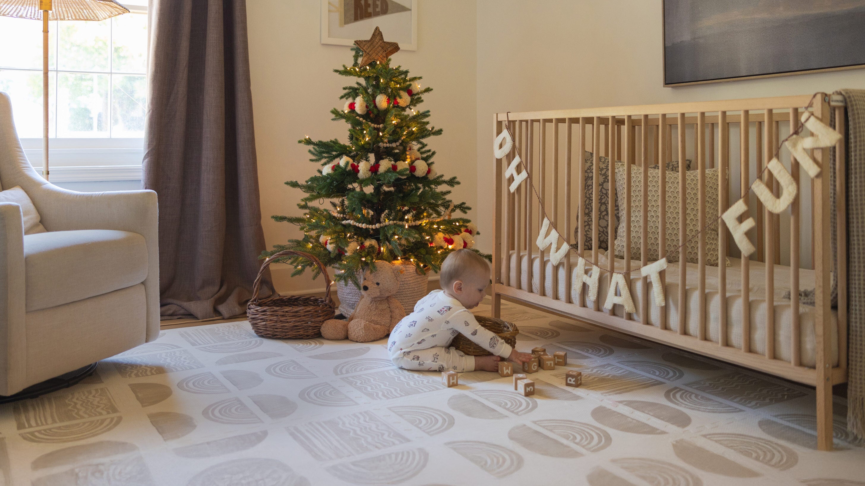 Cream and tan nursery room with a child sitting on a play mat near a crib and Christmas tree.