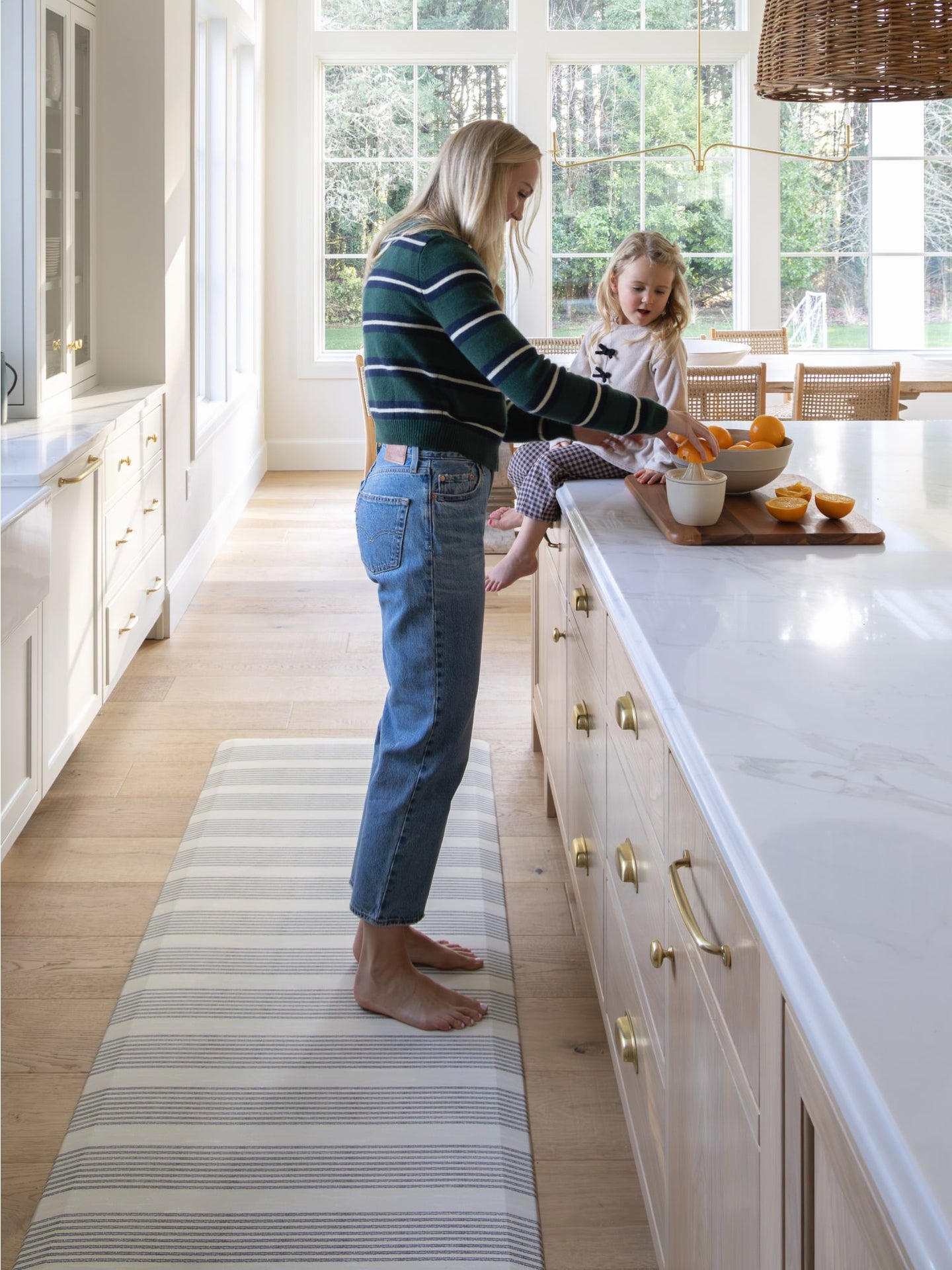 Woman standing on striped kitchen mat juicing oranges on a kitchen counter with toddler sitting on the counter watching her.