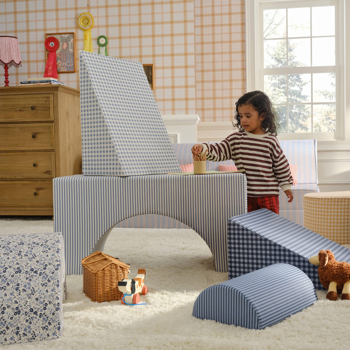 A toddler in a striped sweater plays beside a blue striped arch block topped with a gingham wedge, in a nursery with plaid wallpaper.
